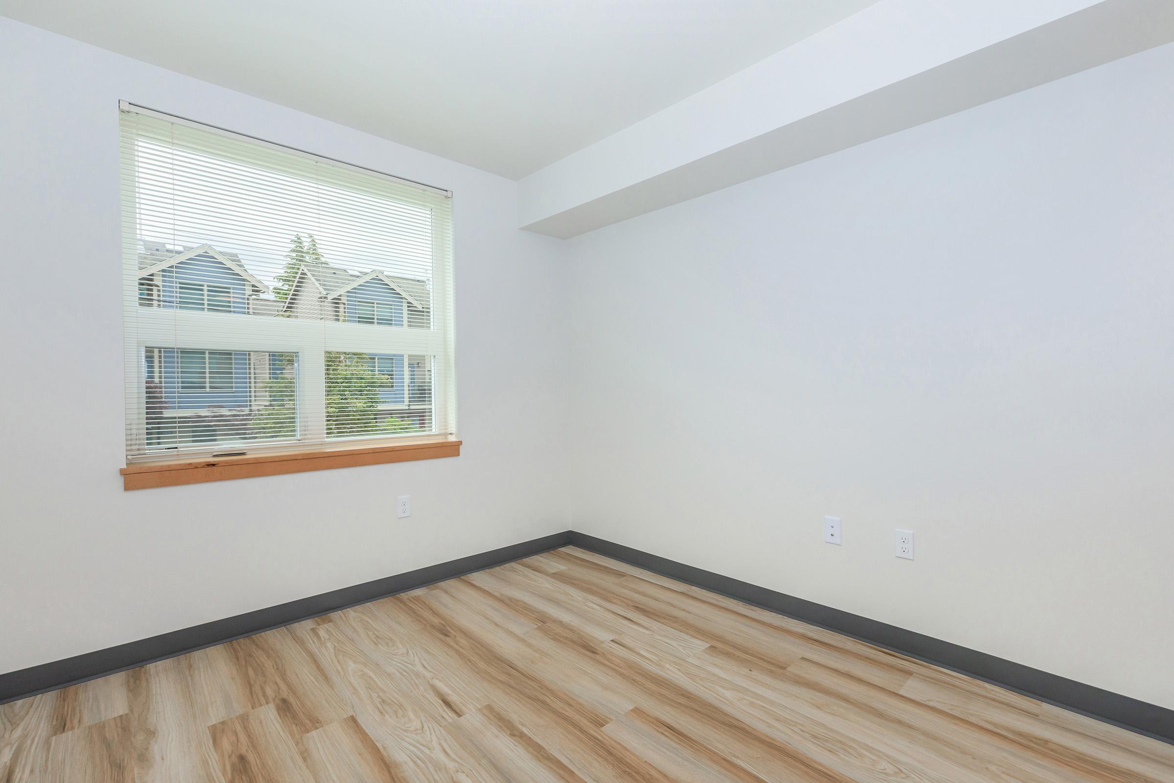 A well-lit, empty room featuring light wood flooring and a large window with blinds. The walls are painted white, and there is a wooden window sill. Outside the window, a view of neighboring houses is visible. The space is unadorned, making it suitable for various uses or furnishings.