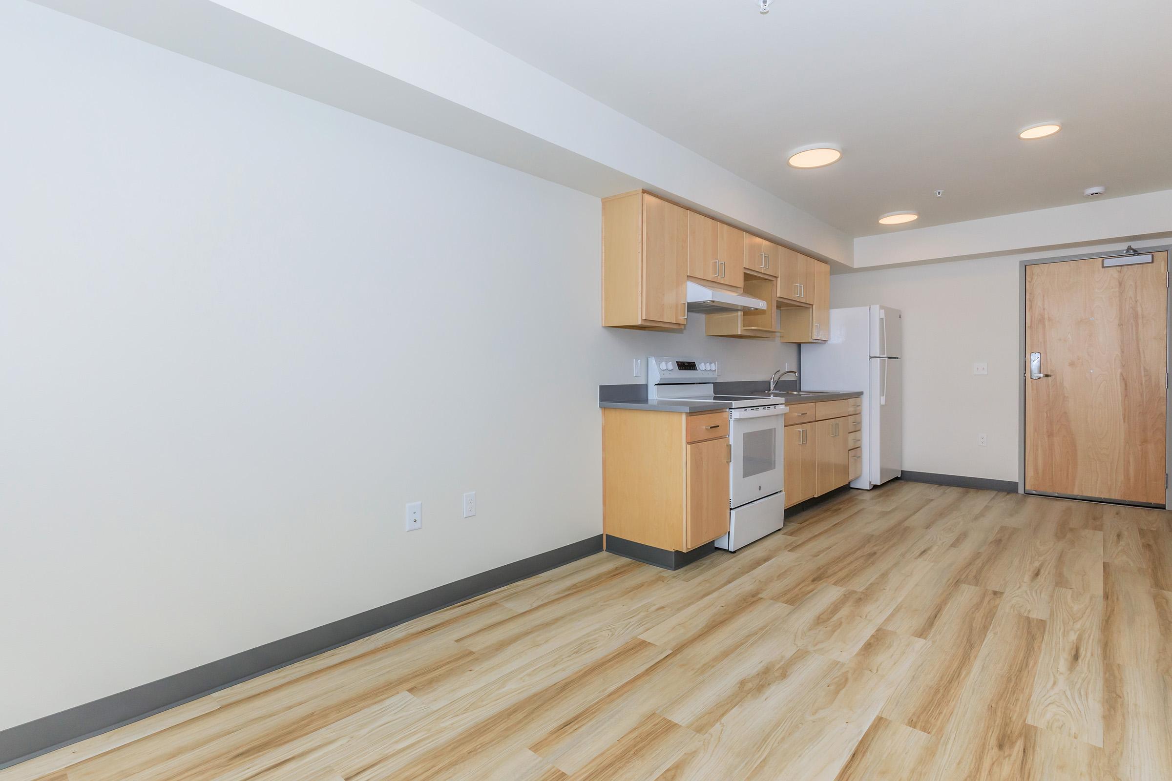 A small, modern kitchen area with light wood cabinetry, a white refrigerator, stove, and microwave. The floor is light-colored wood, and the walls are painted a soft color. The space is well-lit with overhead lights and has a door on the right leading to another room.