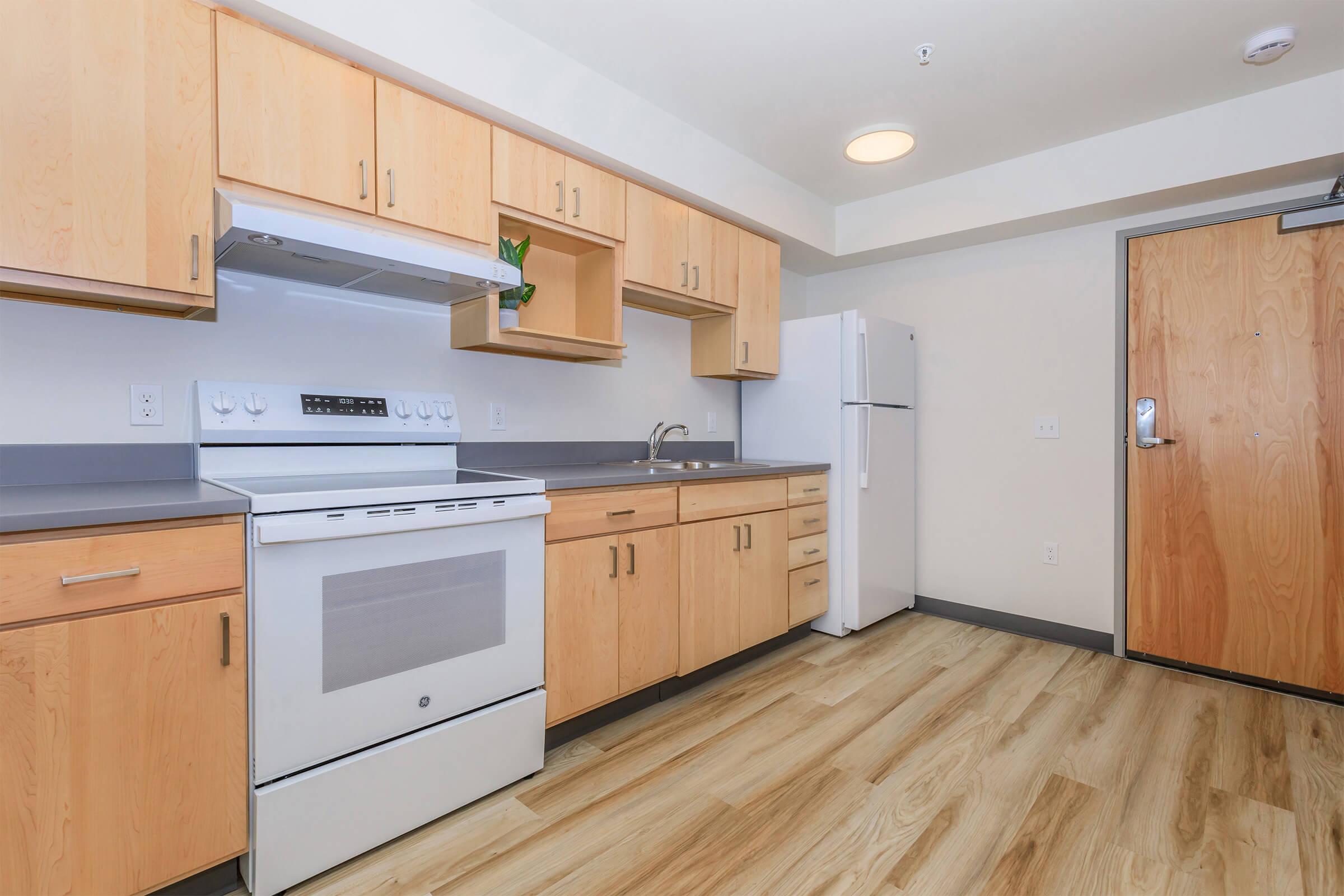 A modern kitchen featuring light wooden cabinetry, a white stove and oven, a stainless steel sink, and a refrigerator. The flooring is light-toned wood, and there are minimal decorations. The kitchen is well-lit with a window and ceiling lights, providing a spacious and clean appearance.