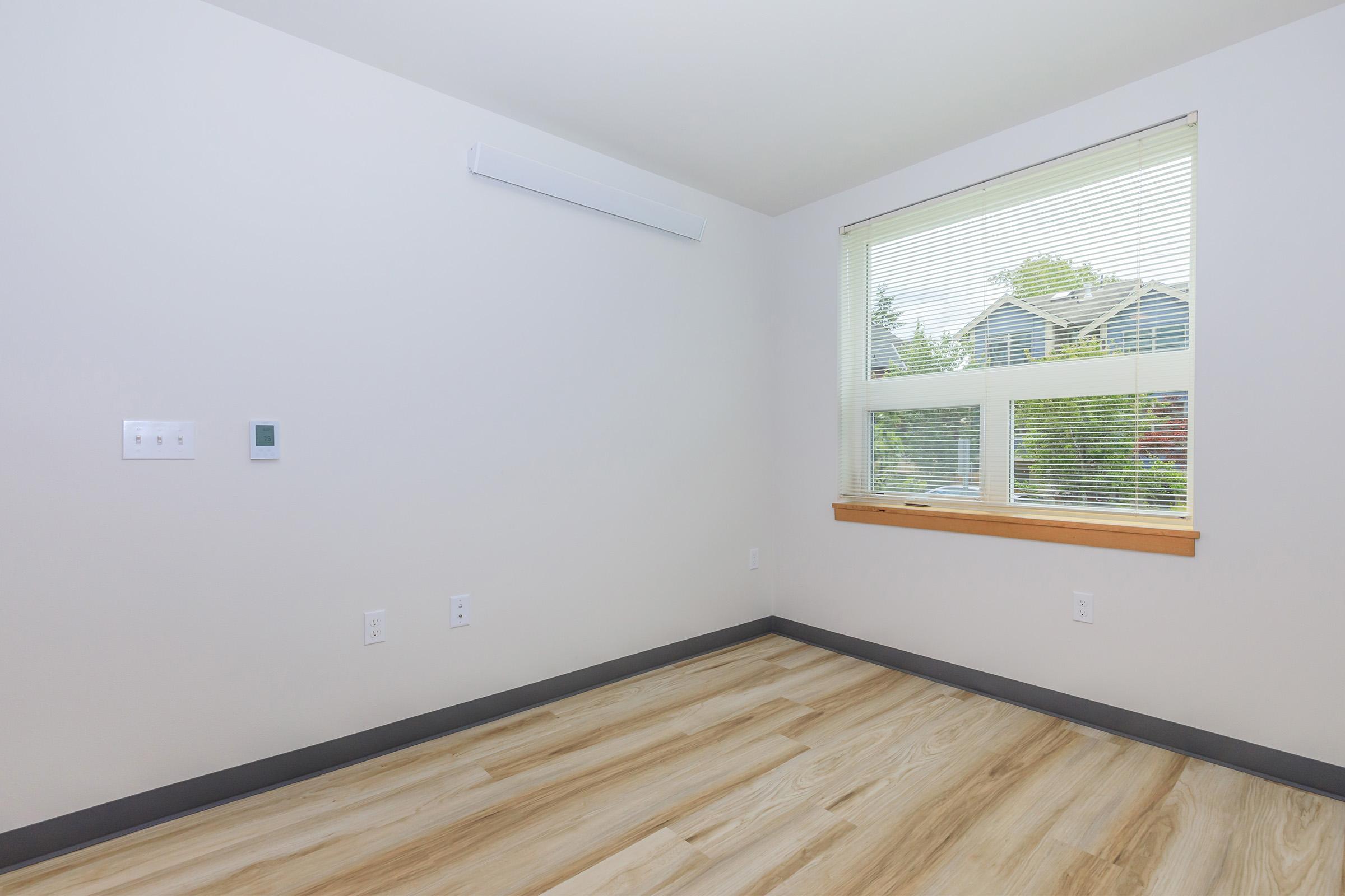 A clean and minimally furnished room featuring a single window with blinds, natural light streaming in, and light-colored wood flooring. The walls are off-white, and there are light switches and a thermostat on one wall. The space has a modern and airy feel.
