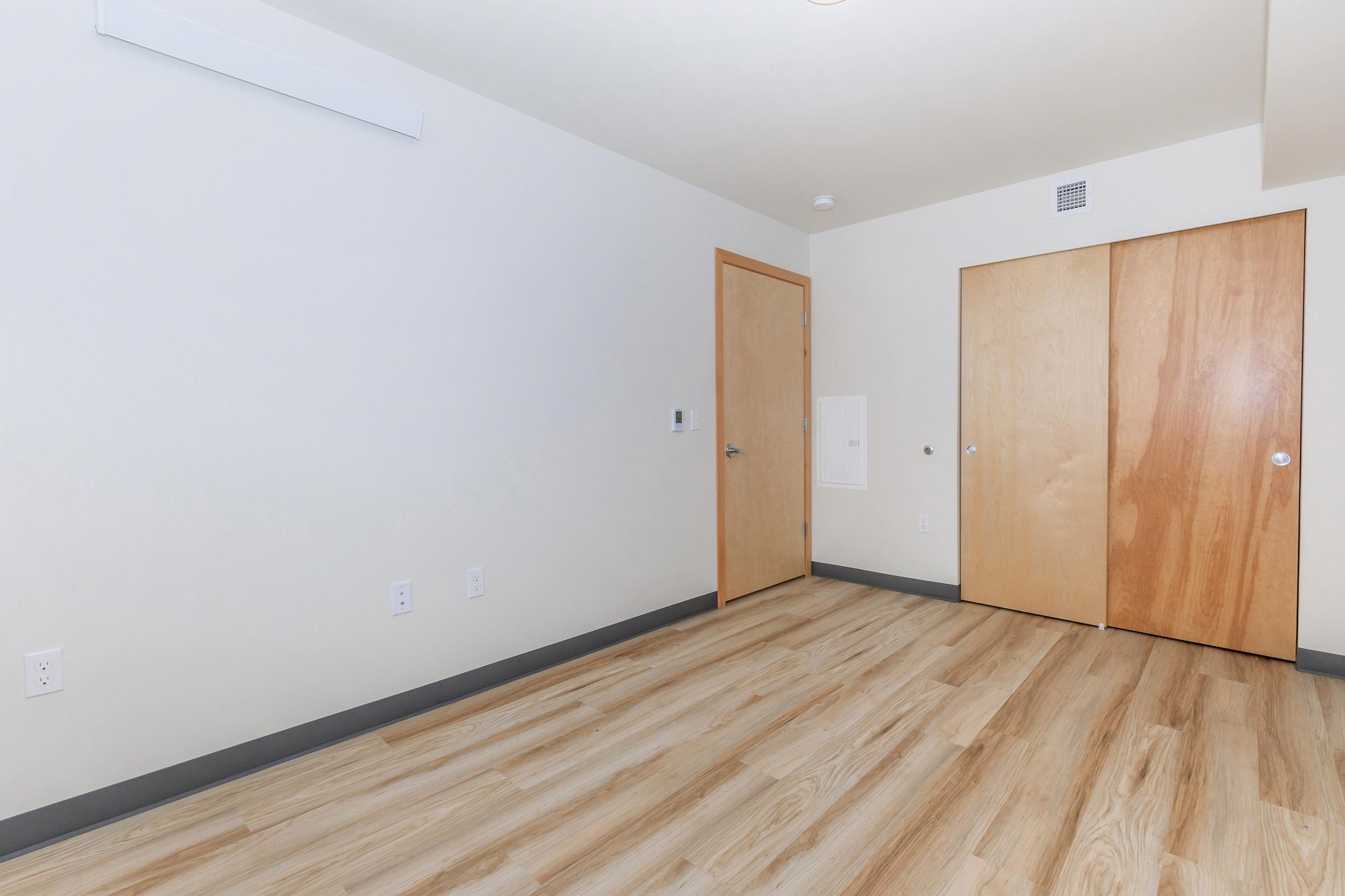 A minimalist interior room featuring light wood flooring, plain white walls, and two wooden doors on the right. The room is empty with no furniture and has a single air vent on the ceiling, creating a clean and spacious appearance.