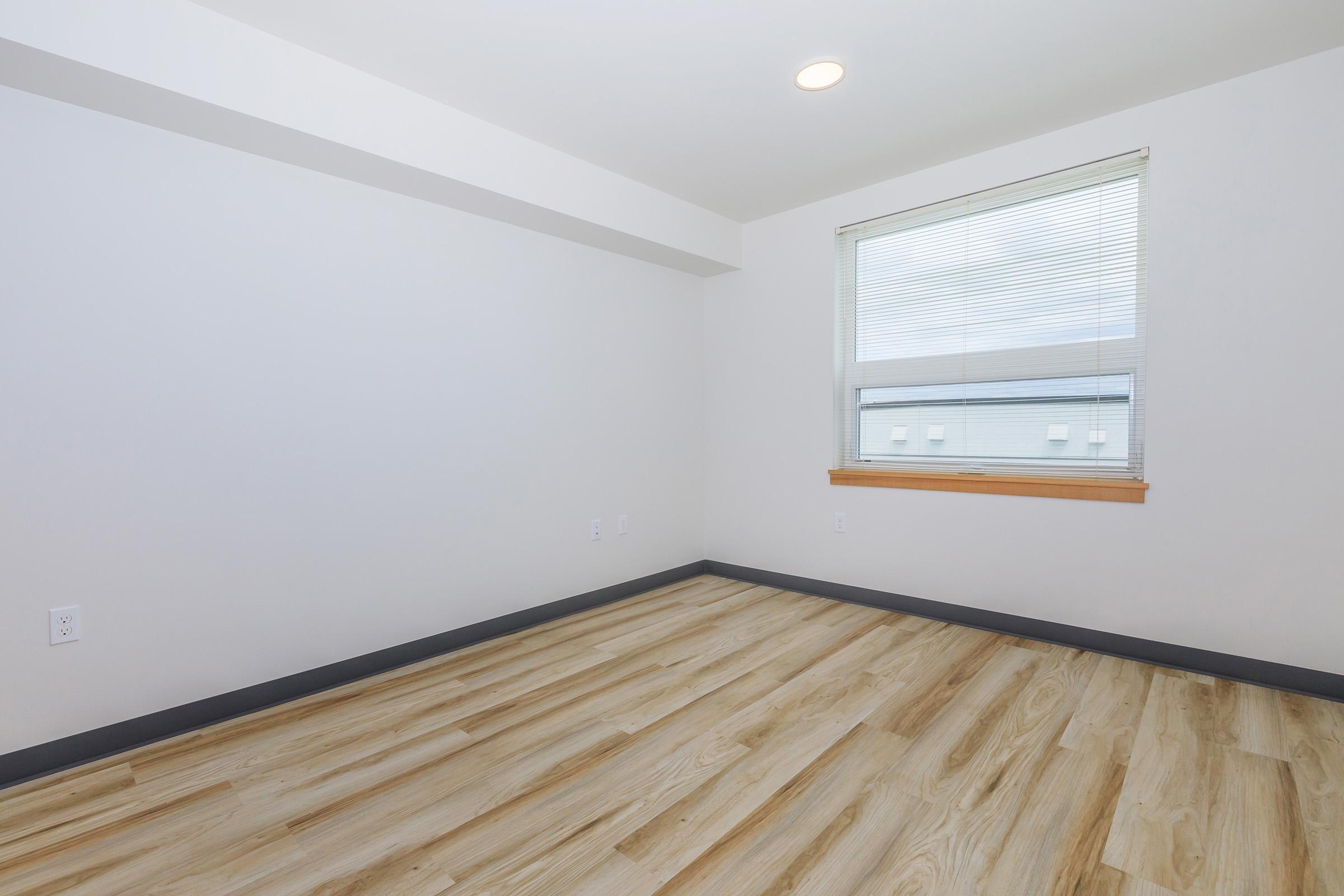 Empty room with light-colored wooden flooring and white walls. A window with blinds is positioned on the far wall, letting in natural light. The room has a simple, modern design with no furniture or decorations.