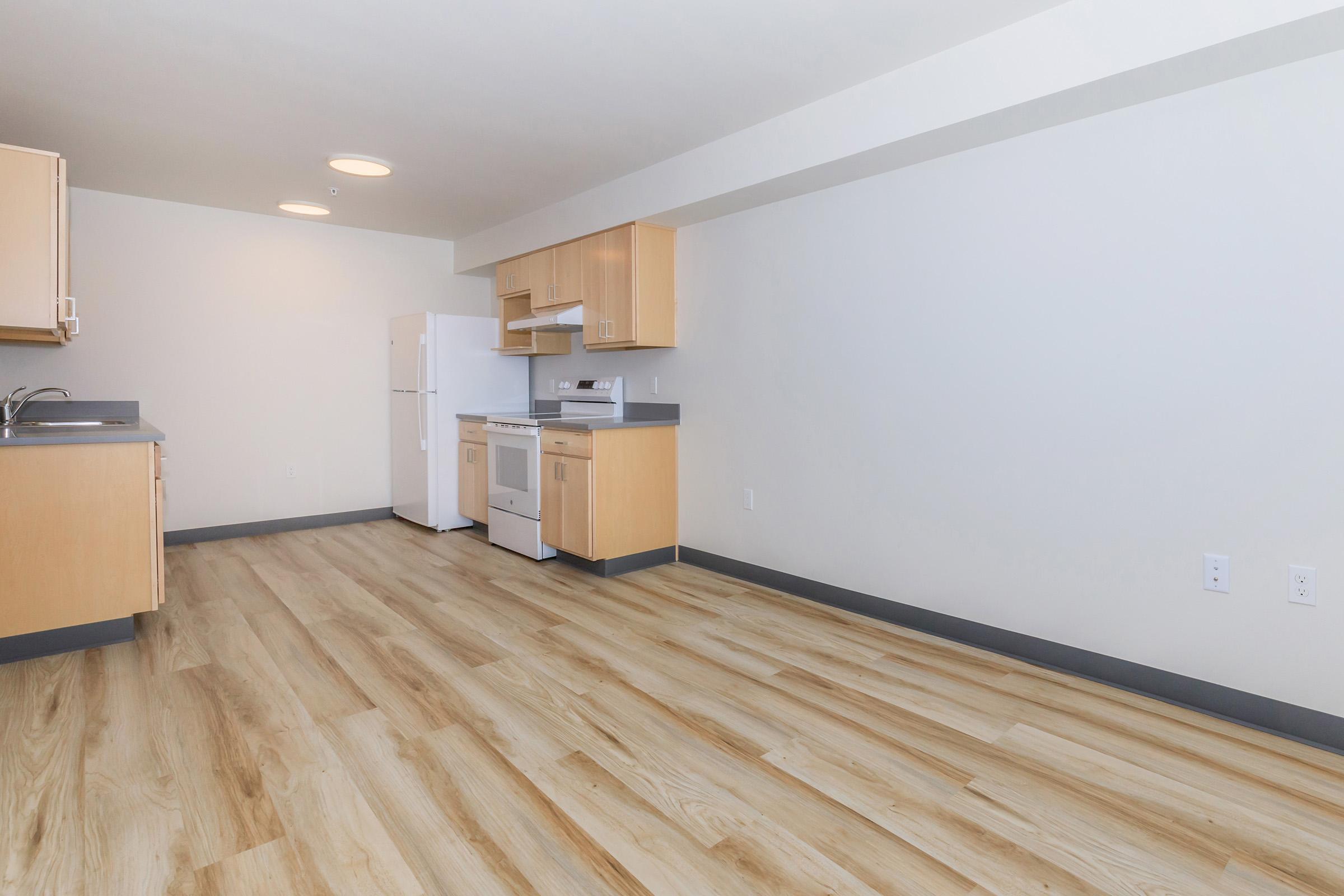 A modern kitchen with light wood cabinetry, a white refrigerator, and a stove. The floor features light wood laminate, and the walls are painted in a neutral tone. The space is well-lit with recessed lighting, creating a clean and open atmosphere.