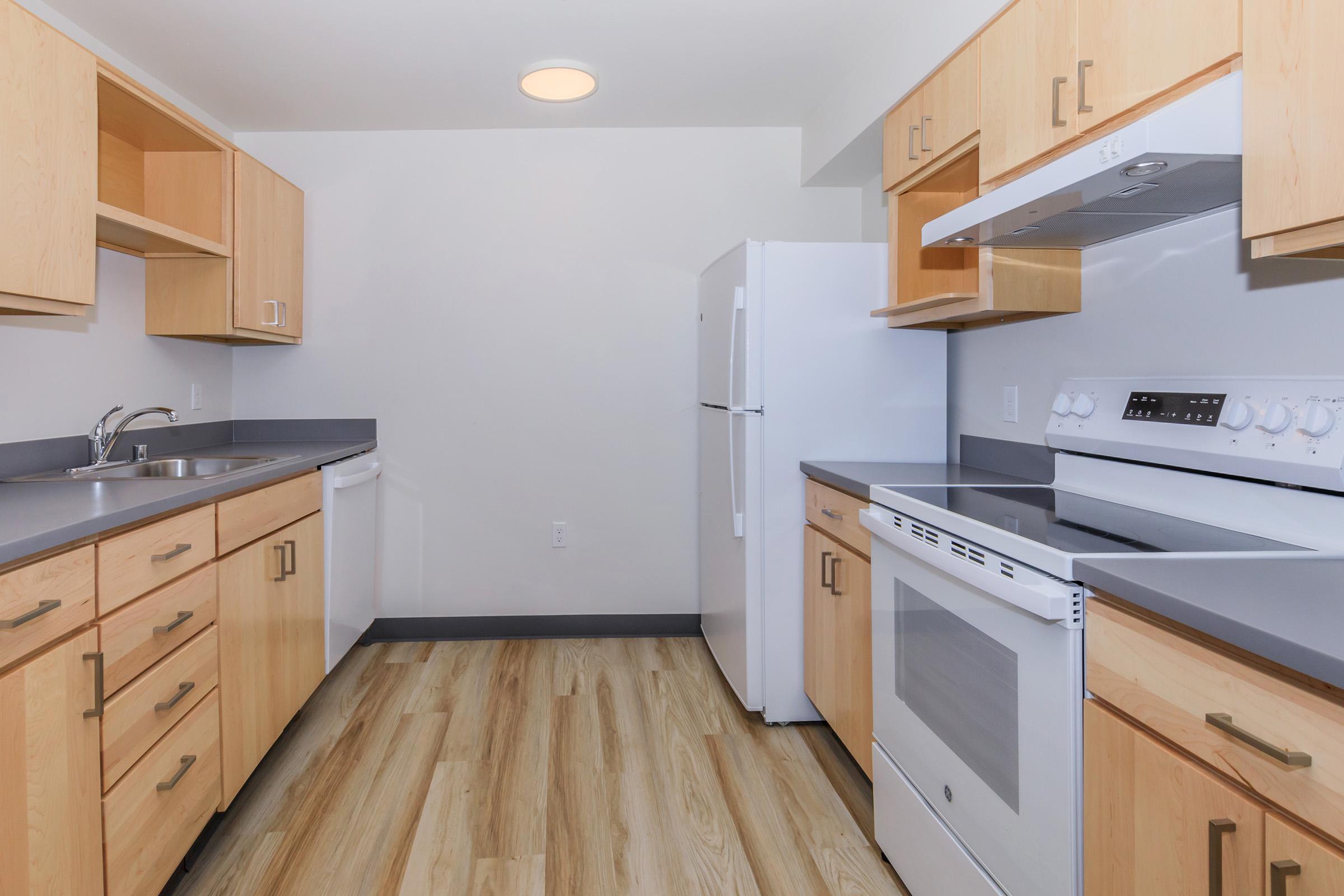 Modern kitchen featuring light wood cabinetry, stainless steel appliances, a white stove and oven, a refrigerator, and a sink. The countertops are gray, and the flooring is a light wood laminate, creating a bright and spacious environment.