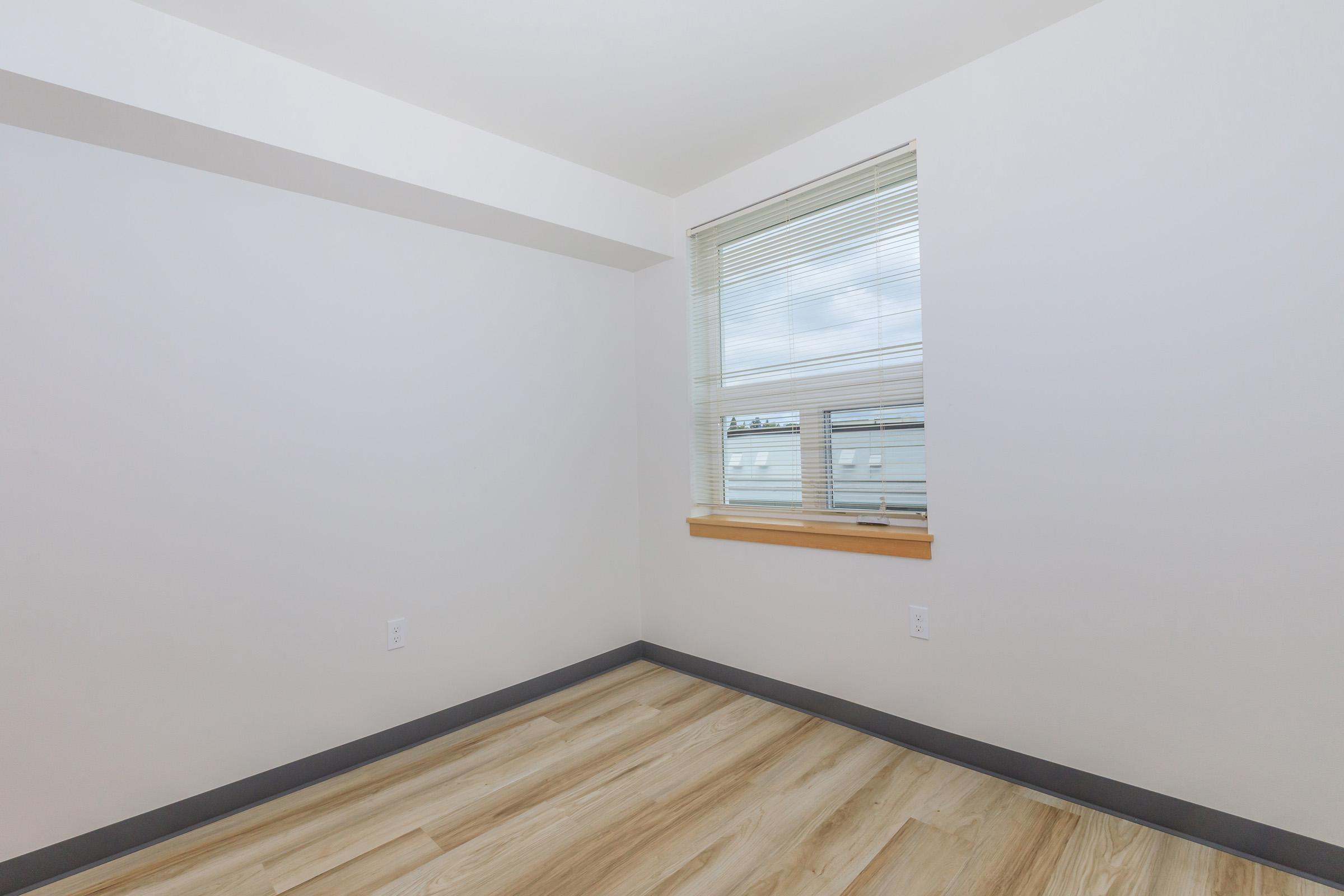 A vacant room with light-colored walls and wooden flooring. There is a single window with blinds, allowing natural light to enter. The space appears clean, simple, and ready for decoration or furnishing.