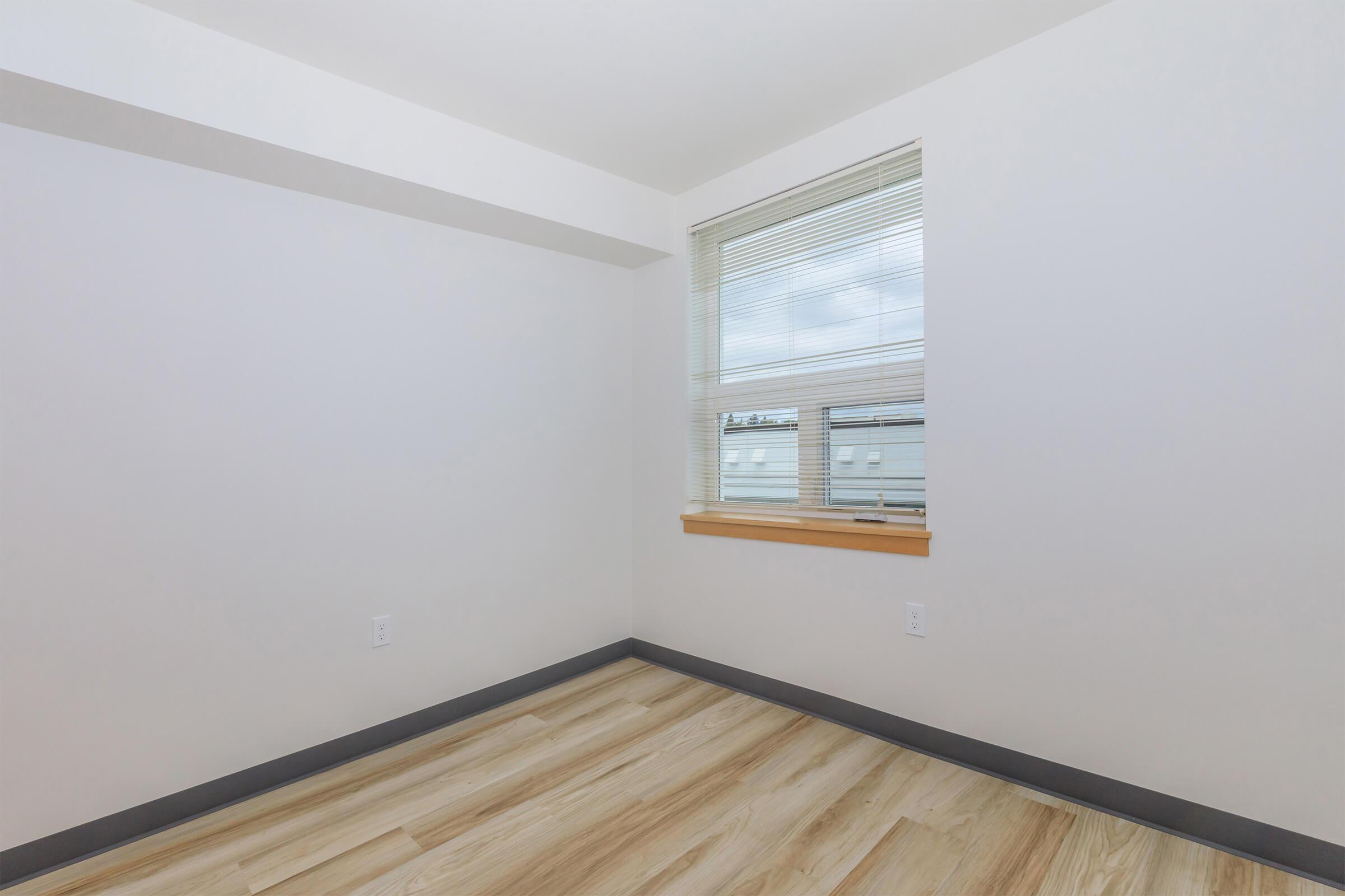 A bright, empty room with light wood flooring and white walls. A window with blinds provides natural light, and there are no furnishings or decorations present. The corner of the room is visible, emphasizing the spacious, minimalist design.