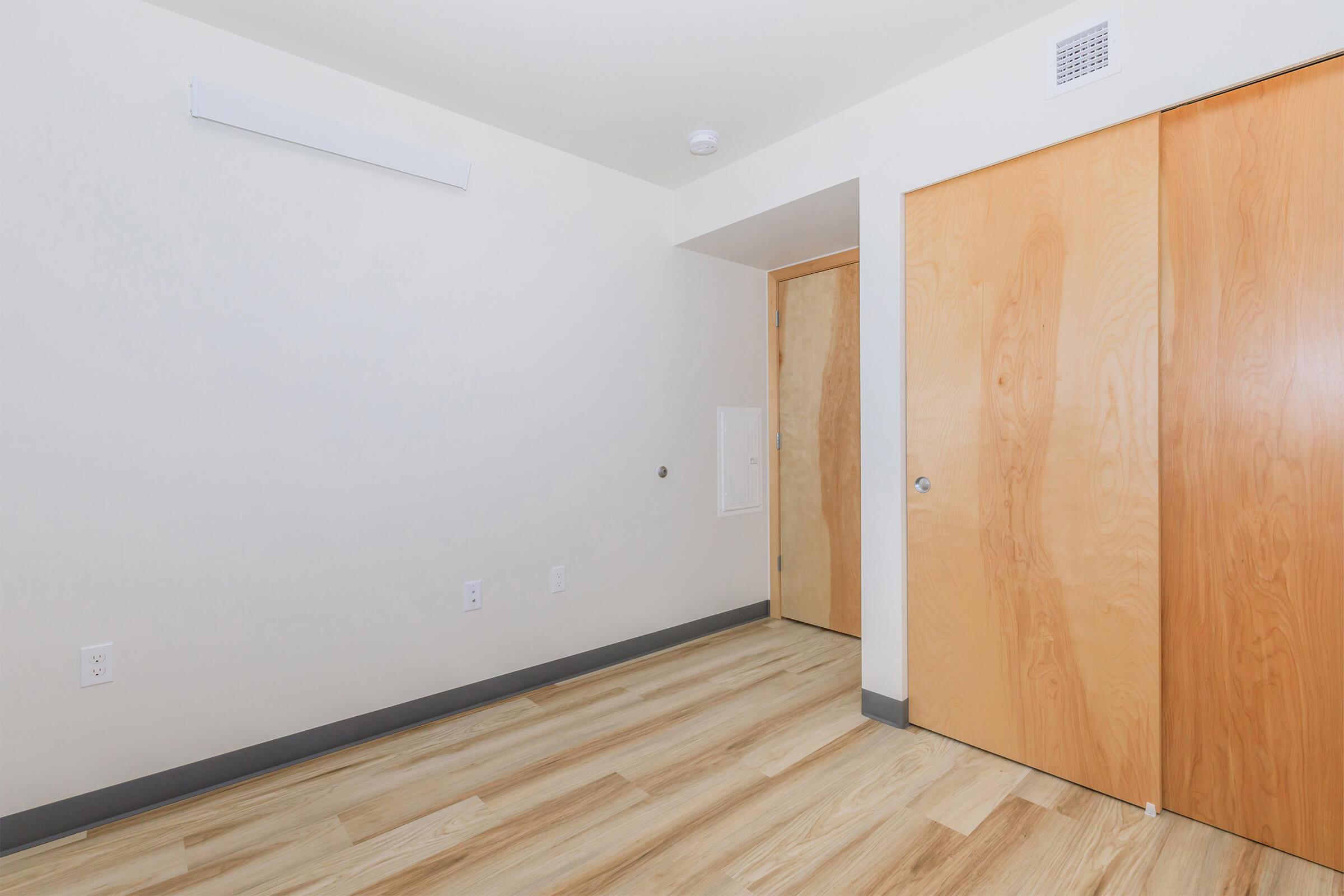 A well-lit, empty room with light wood flooring, featuring a white wall and two wooden doors. One door is partially open, leading to another area. There’s a vent on the ceiling and no furniture present, creating a spacious and minimalist appearance.