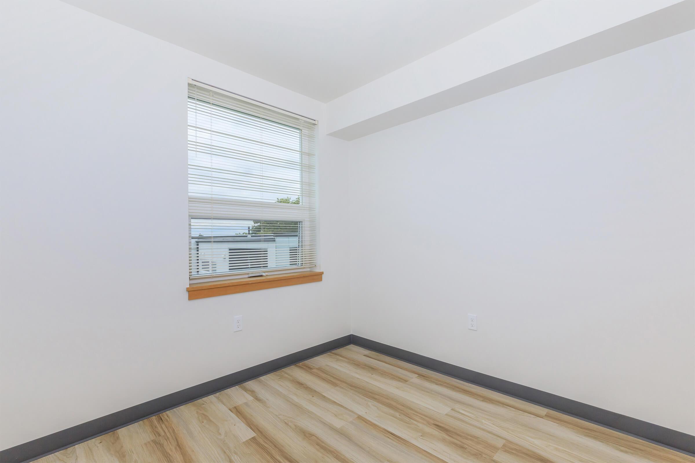 An empty room with a light-colored wooden floor, white walls, and a window featuring blinds. The window allows natural light to enter, and a small ledge is visible beneath it. The overall atmosphere is bright and minimalist, creating a spacious feel.