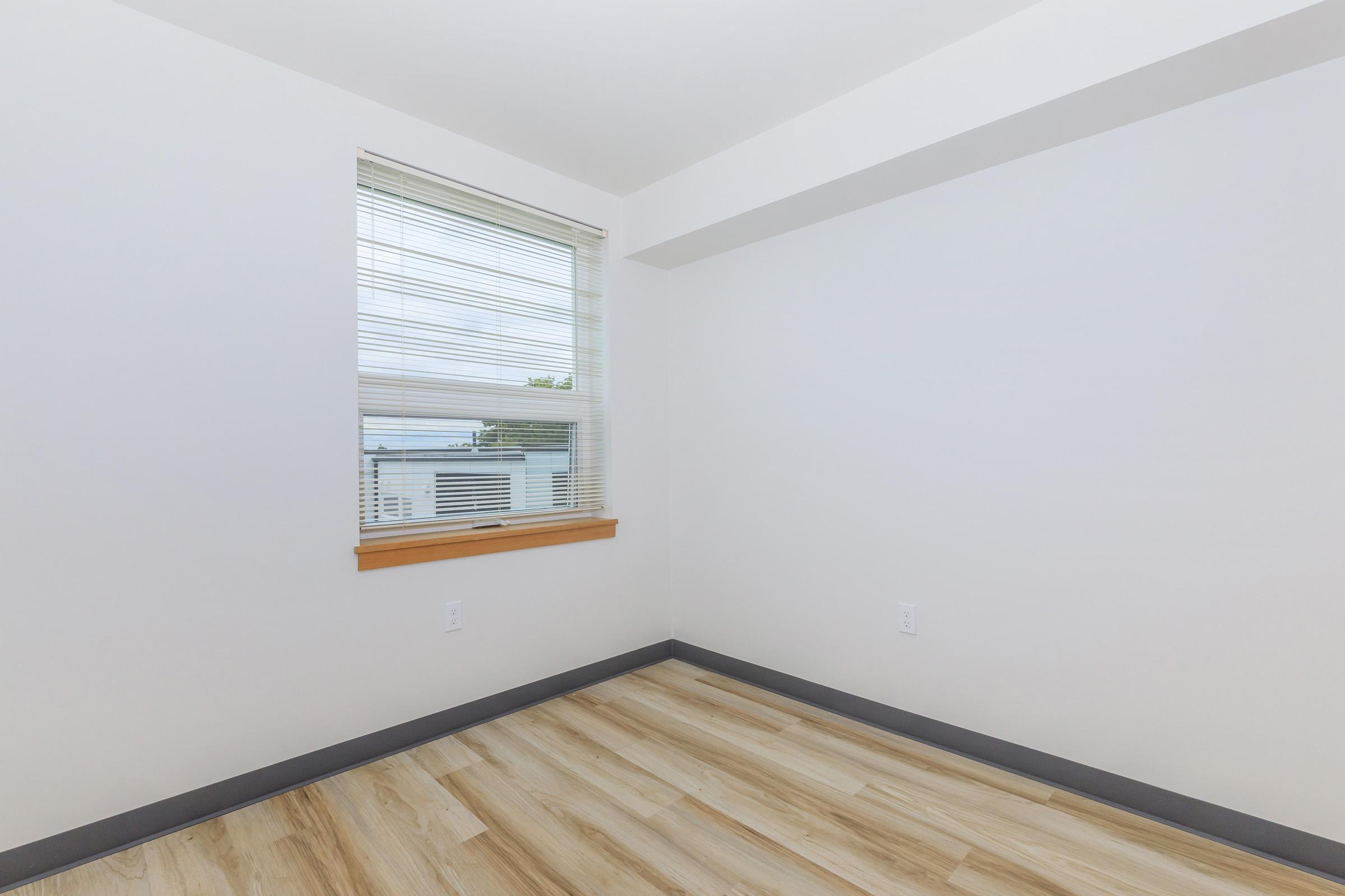 A bright, empty room with light-colored walls and a hardwood floor. A window with blinds allows natural light to enter, and there is a small wooden ledge below the window. The space has a modern and minimalistic feel, with no furniture or decorations present.