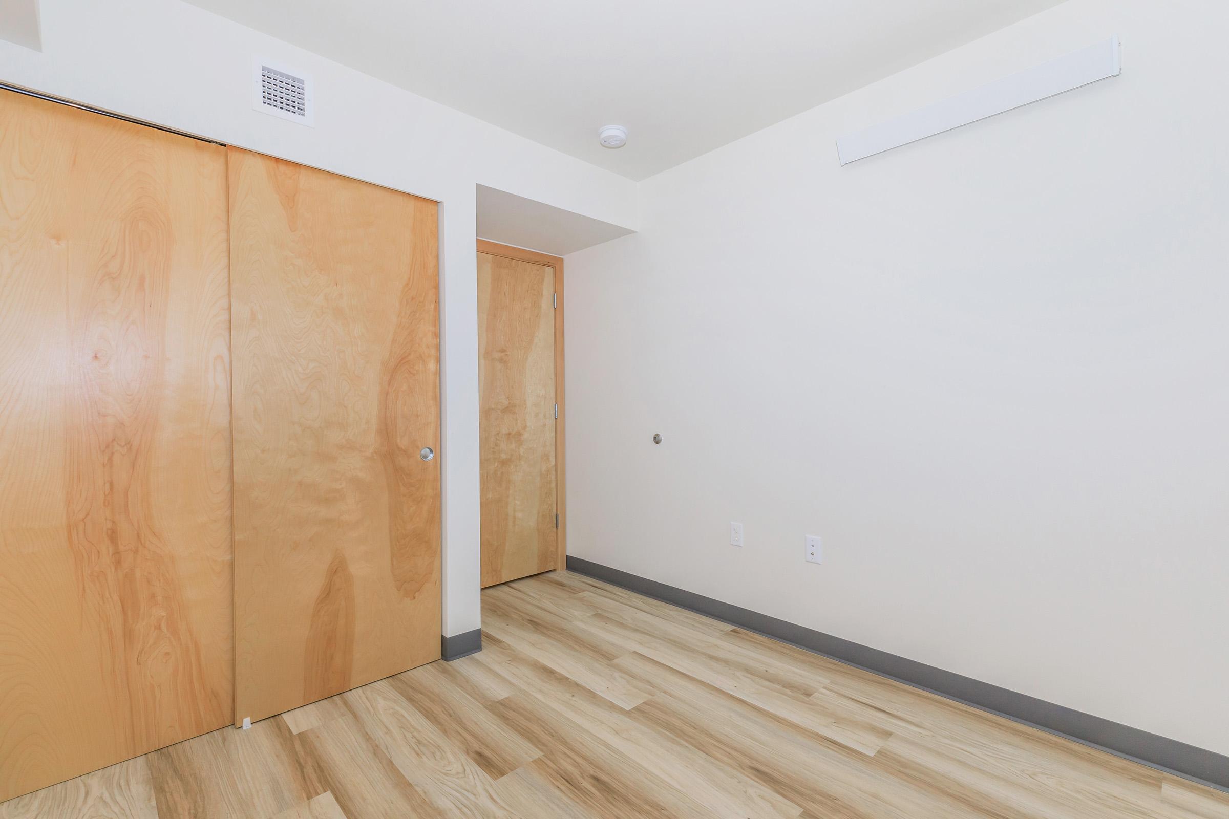 Interior view of a room featuring light-colored wooden flooring, two closed wooden doors, and plain white walls. The space is minimalistic with a bright and airy feel, lacking furniture or decorations. A small ceiling vent is visible, contributing to the modern design of the room.