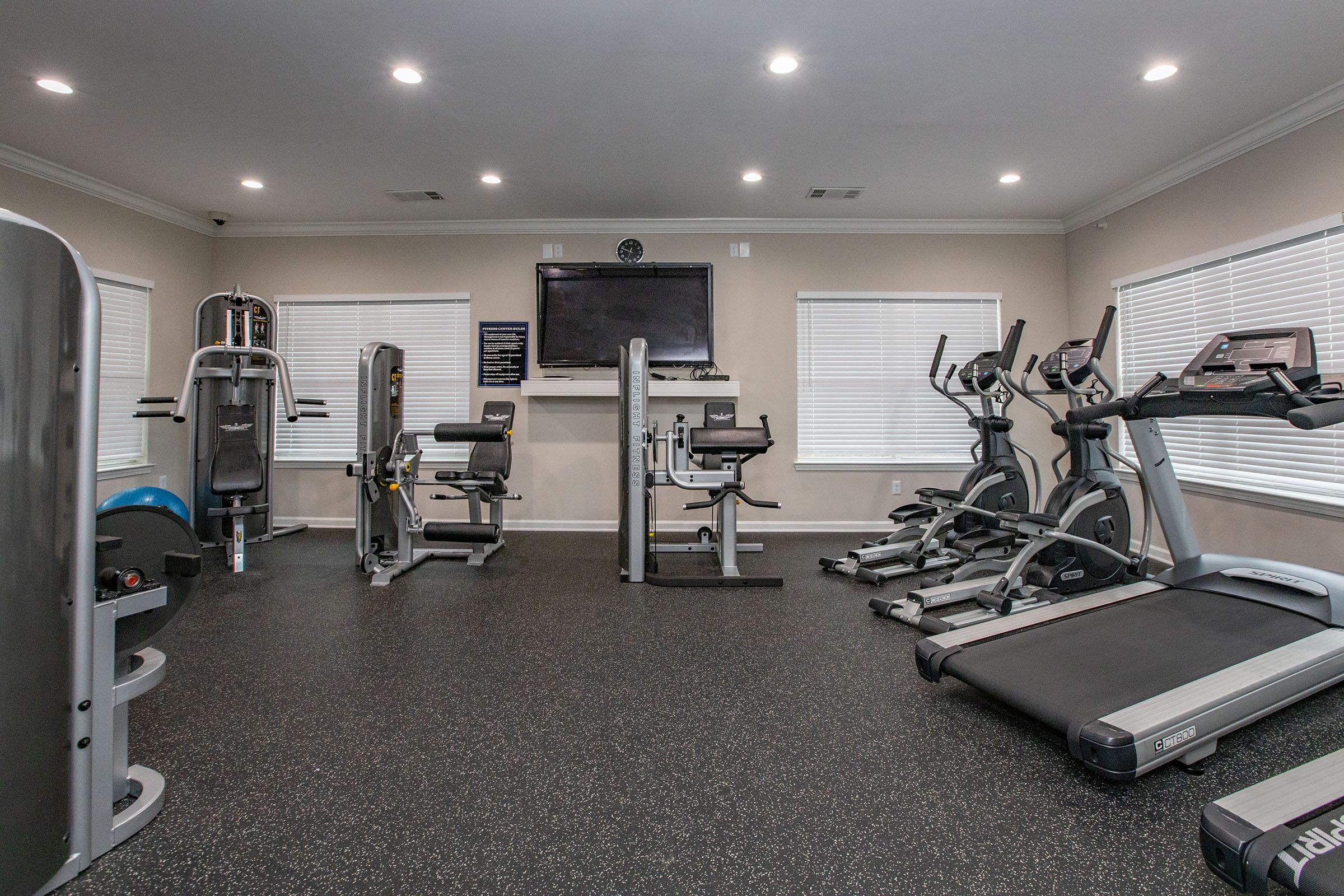 Interior of a modern gym featuring various exercise equipment including treadmills, stationary bikes, and weight machines. The space is well-lit with overhead lights and has windows with white blinds. A television is mounted on the wall.