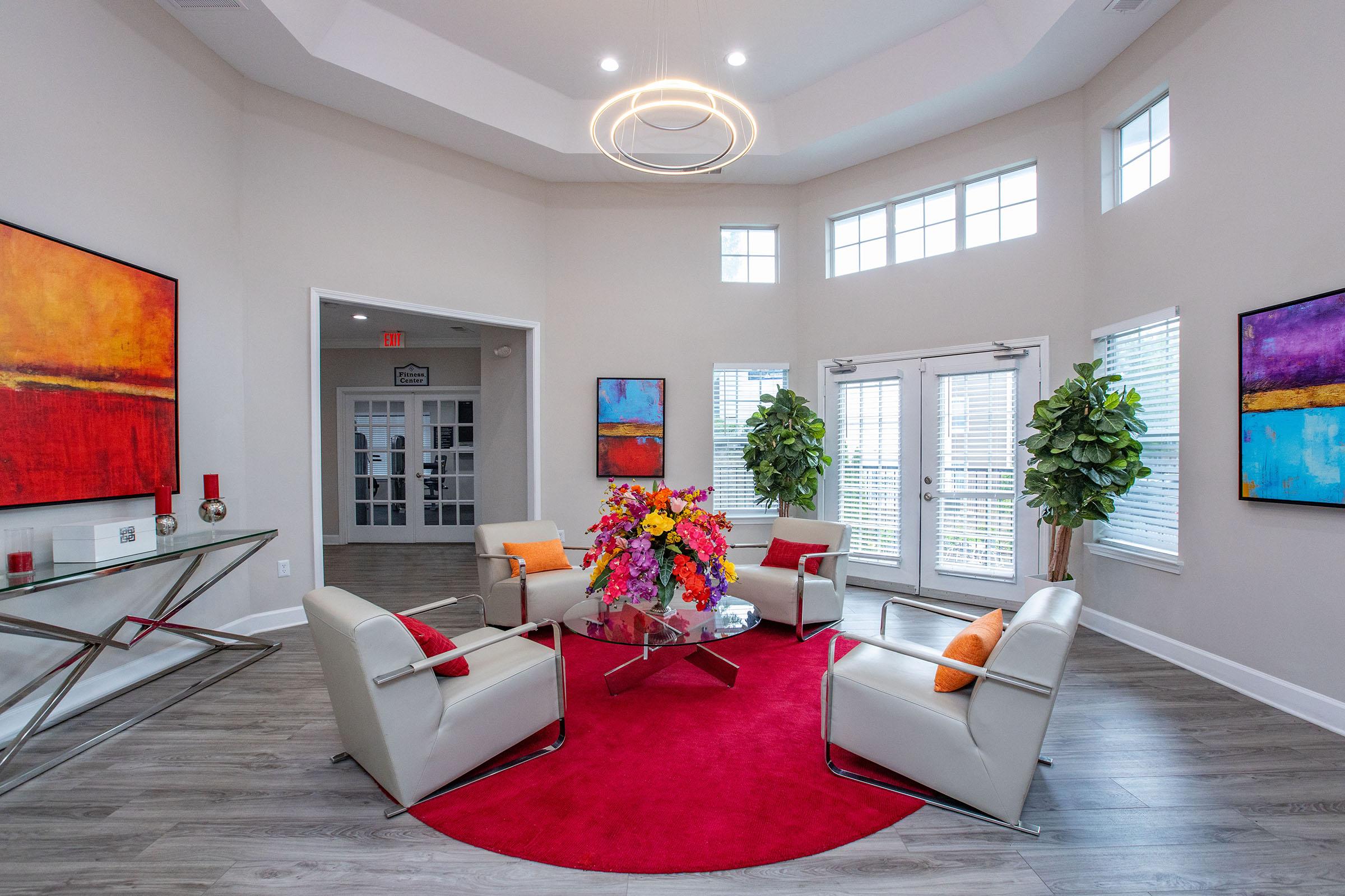 A modern, bright lobby area featuring four contemporary white chairs arranged around a glass coffee table with a vibrant floral centerpiece. The floor is covered with a circular red rug, and two large colorful abstract paintings adorn the walls. Large windows allow natural light to fill the space, and potted plants add a touch of greenery.