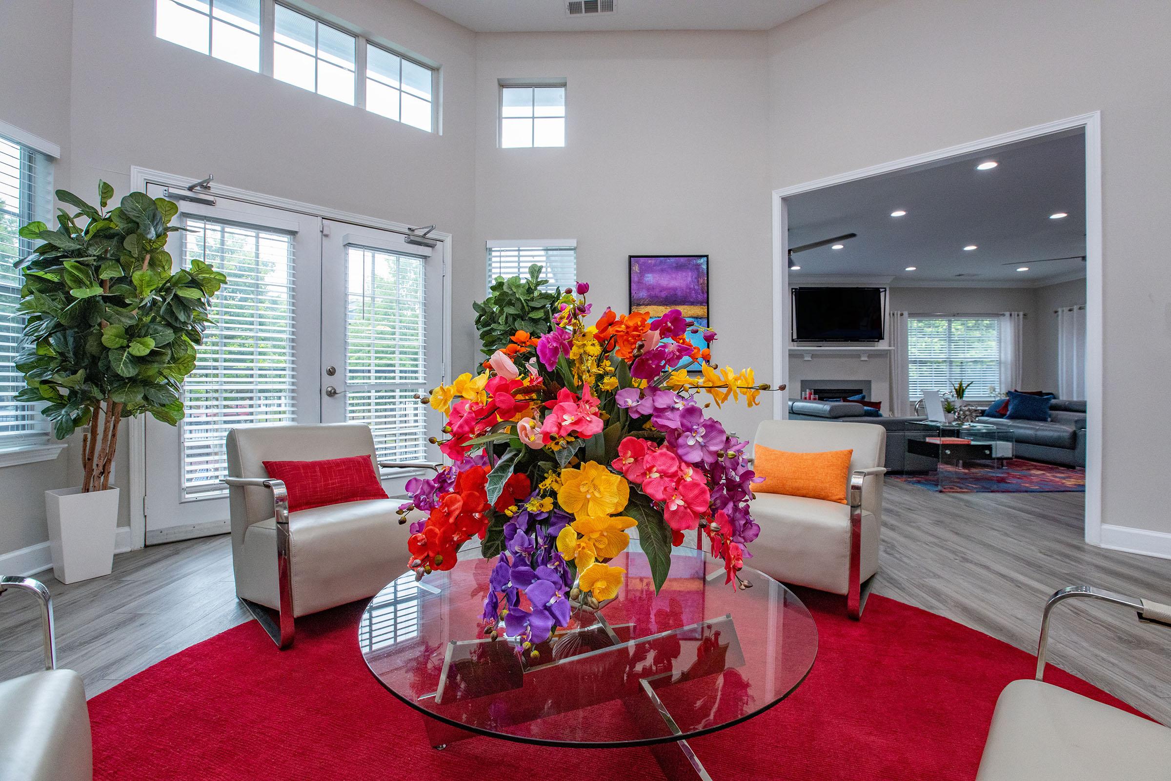 A vibrant indoor living space featuring a glass coffee table with a colorful flower arrangement, two modern chairs, a red area rug, and large windows allowing natural light. The background shows an open living area with a couch and TV.