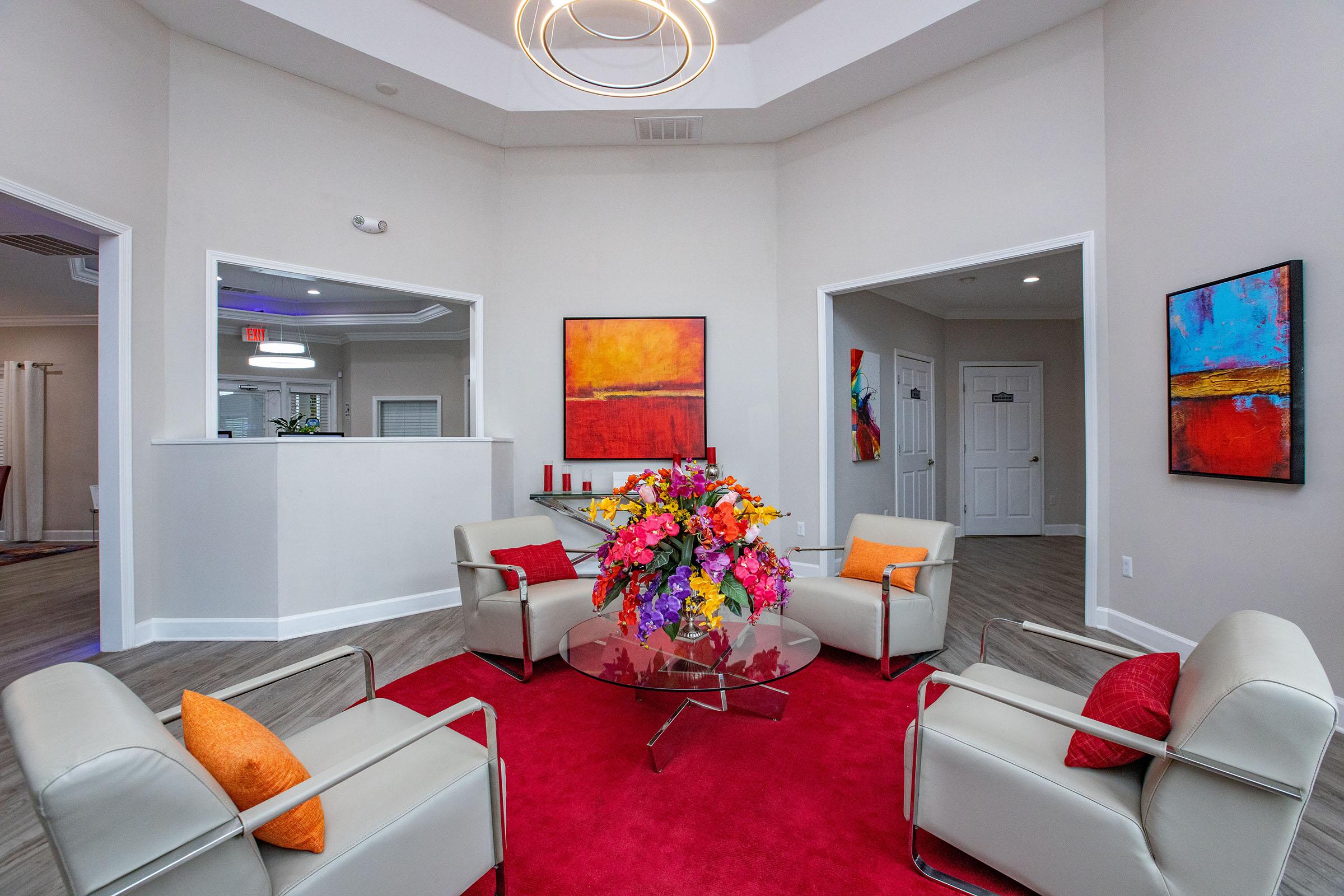 A modern lobby area featuring a circular glass coffee table with a vibrant floral arrangement. Surrounding the table are four light-colored armchairs with orange and red accent pillows. The walls are painted in neutral tones, adorned with colorful abstract artwork. A red area rug adds warmth to the space, which is well-lit and inviting.