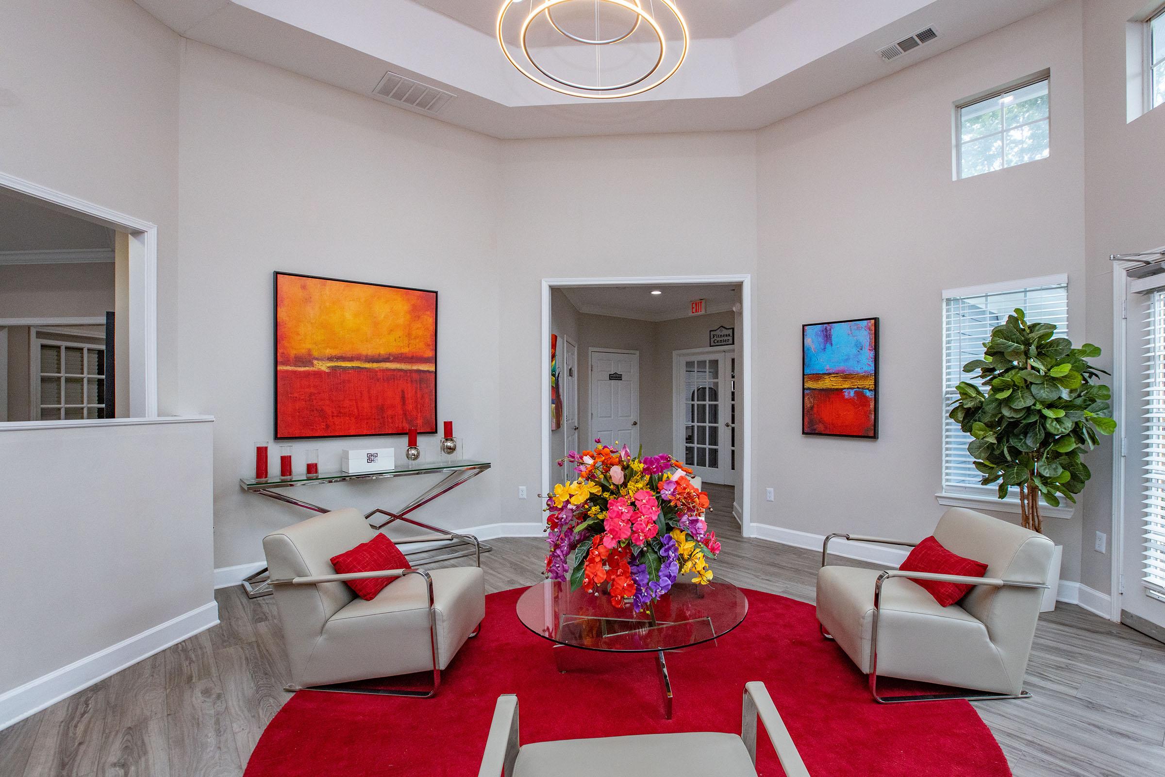 A modern living room featuring two light-colored chairs facing a round glass coffee table with a large floral arrangement. The room has light-colored walls and wood-like flooring, with vibrant abstract paintings on the walls. A red rug adds color to the space, and there are tall windows allowing natural light in, along with a potted plant in the corner.