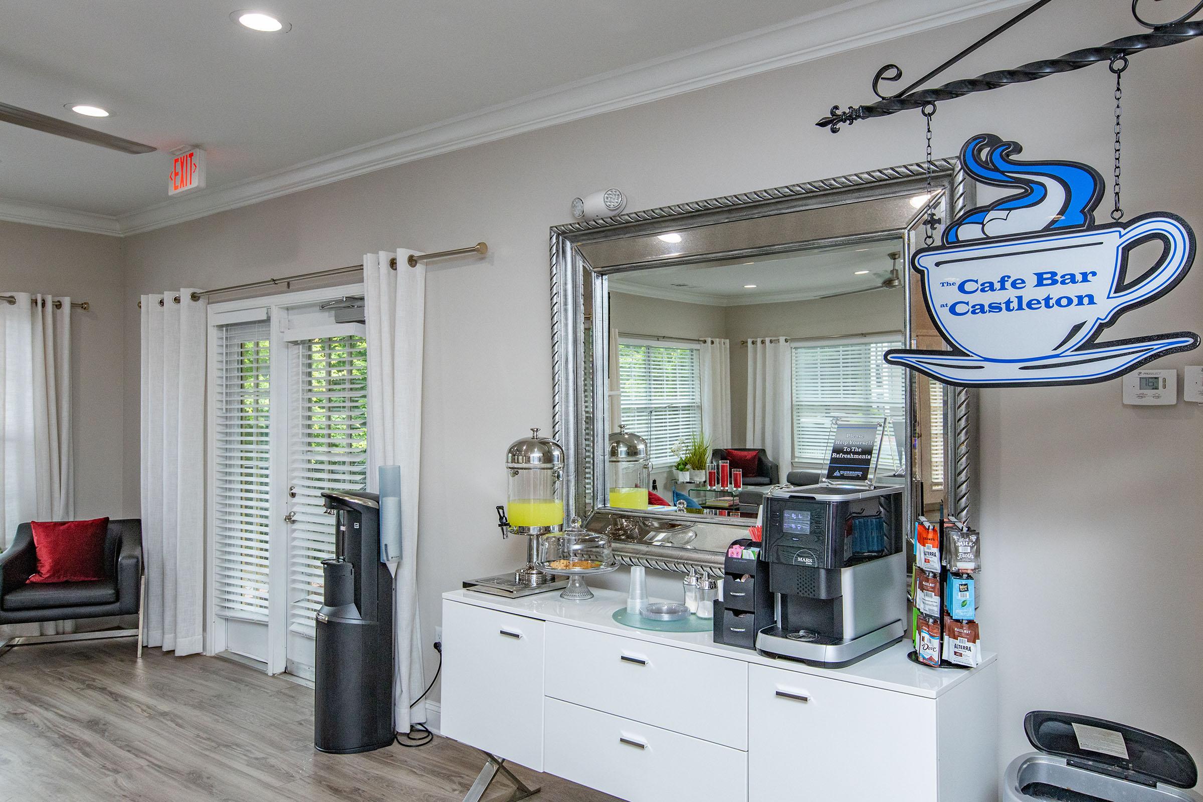 A cafe bar area featuring a decorative sign that says "Cafe Bar Castleton," a coffee machine, a water dispenser, and a mirror reflecting the room. The space has light-colored walls, floor-to-ceiling windows with white blinds, and a cozy seating area with a black armchair and red cushions.