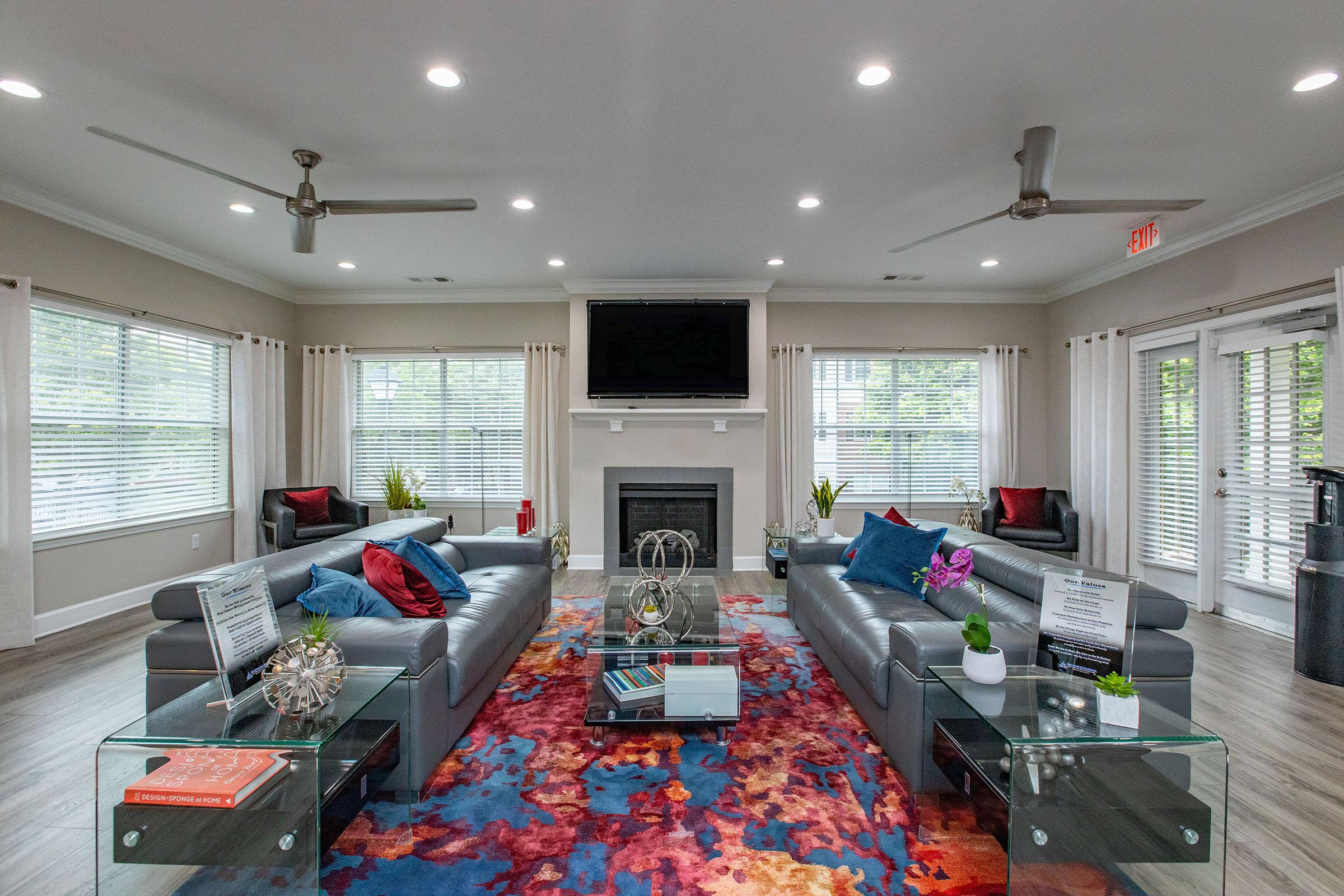 A modern living room featuring two gray sofas with colorful accent pillows, a coffee table with decorative items, and a large area rug displaying vibrant colors. There is a fireplace with a flat-screen TV above it, large windows with white curtains, and potted plants in the corners. The room is well-lit with ceiling lights and has a contemporary design.