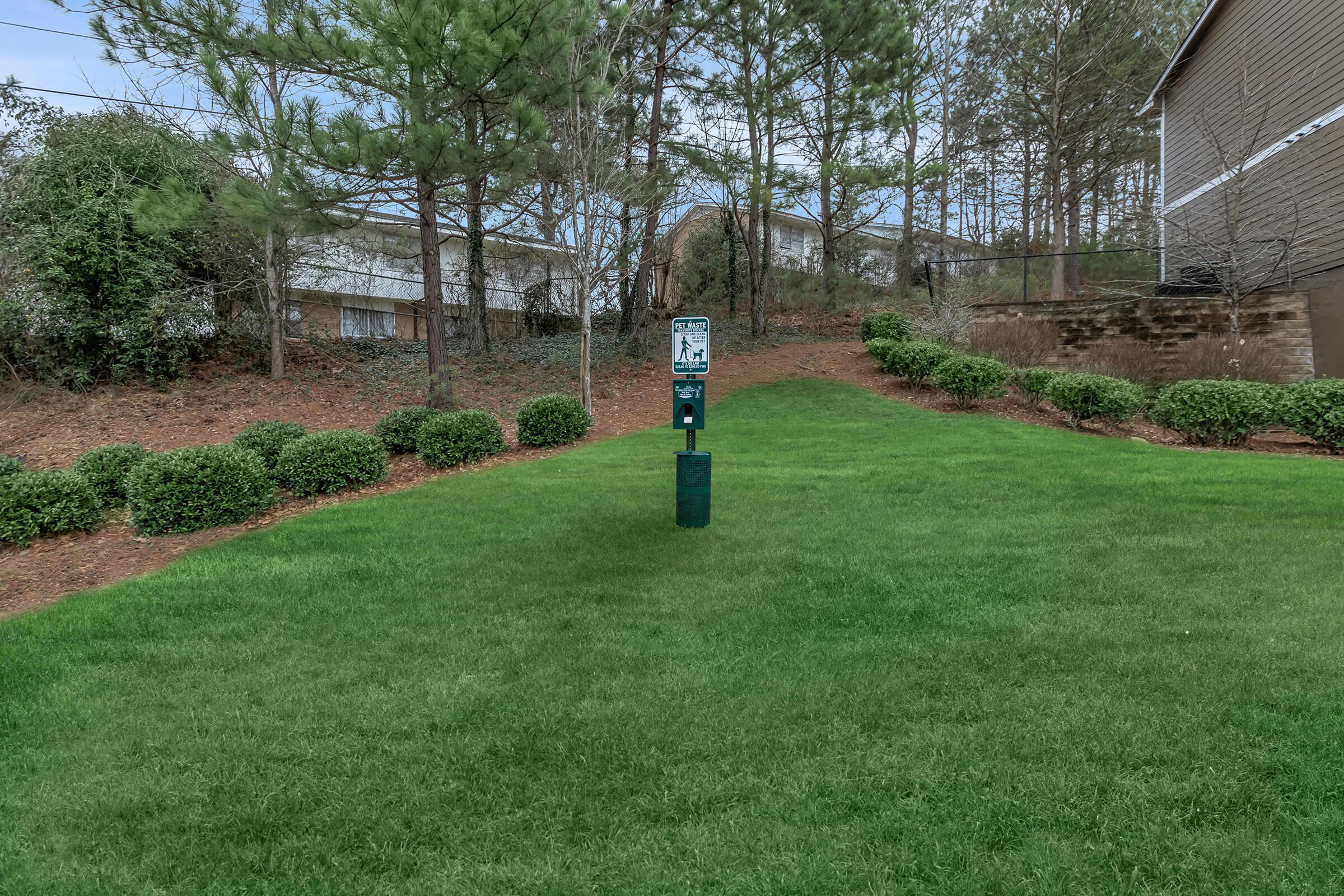 A green lawn with neatly trimmed bushes and a grassy slope, featuring a dog waste station with a sign and bags for pet owners. In the background, there are trees and buildings partially visible.