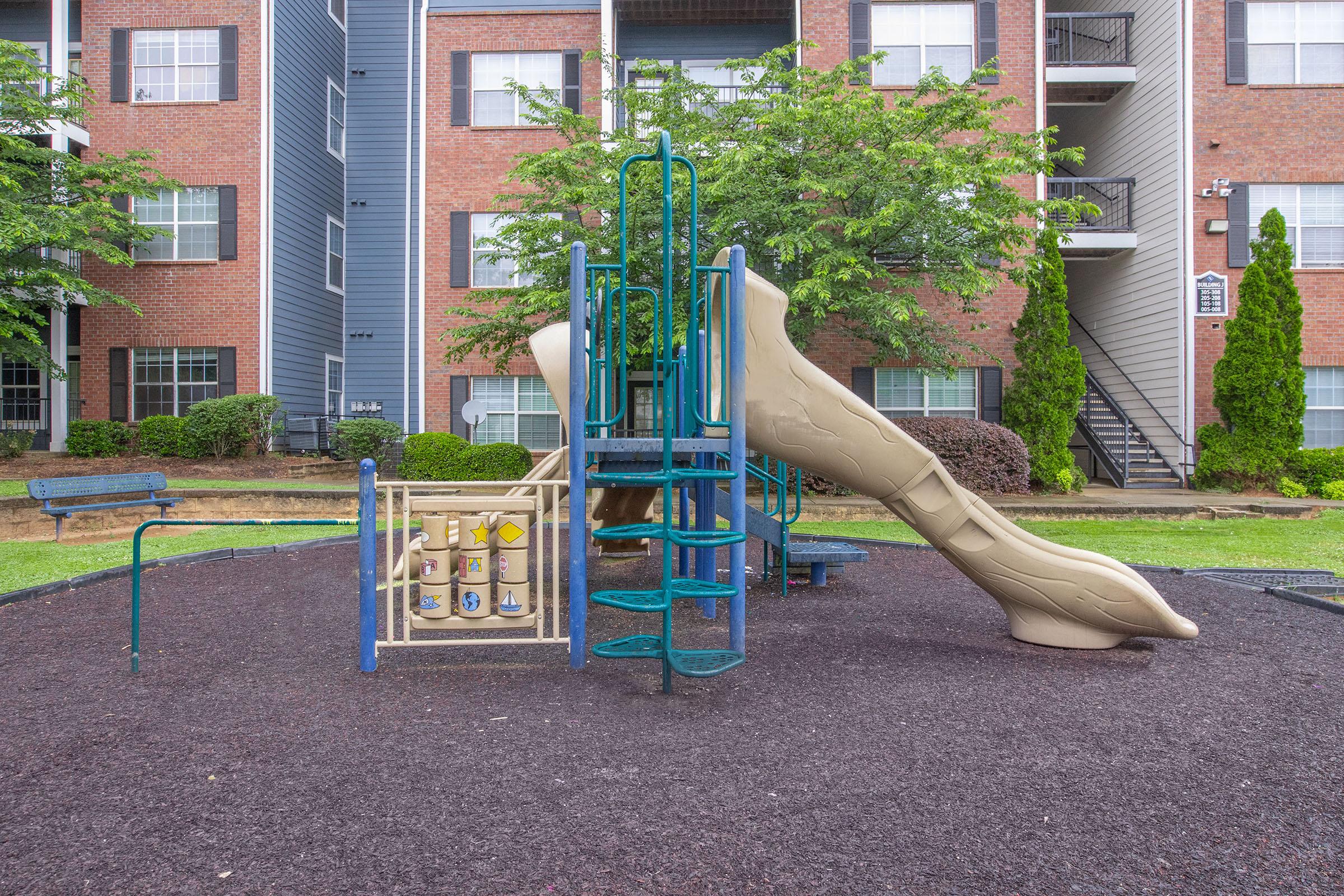 A children's playground featuring a slide, climbing structure, and play panel, situated on a rubber surface surrounded by a grassy area and residential buildings in the background.