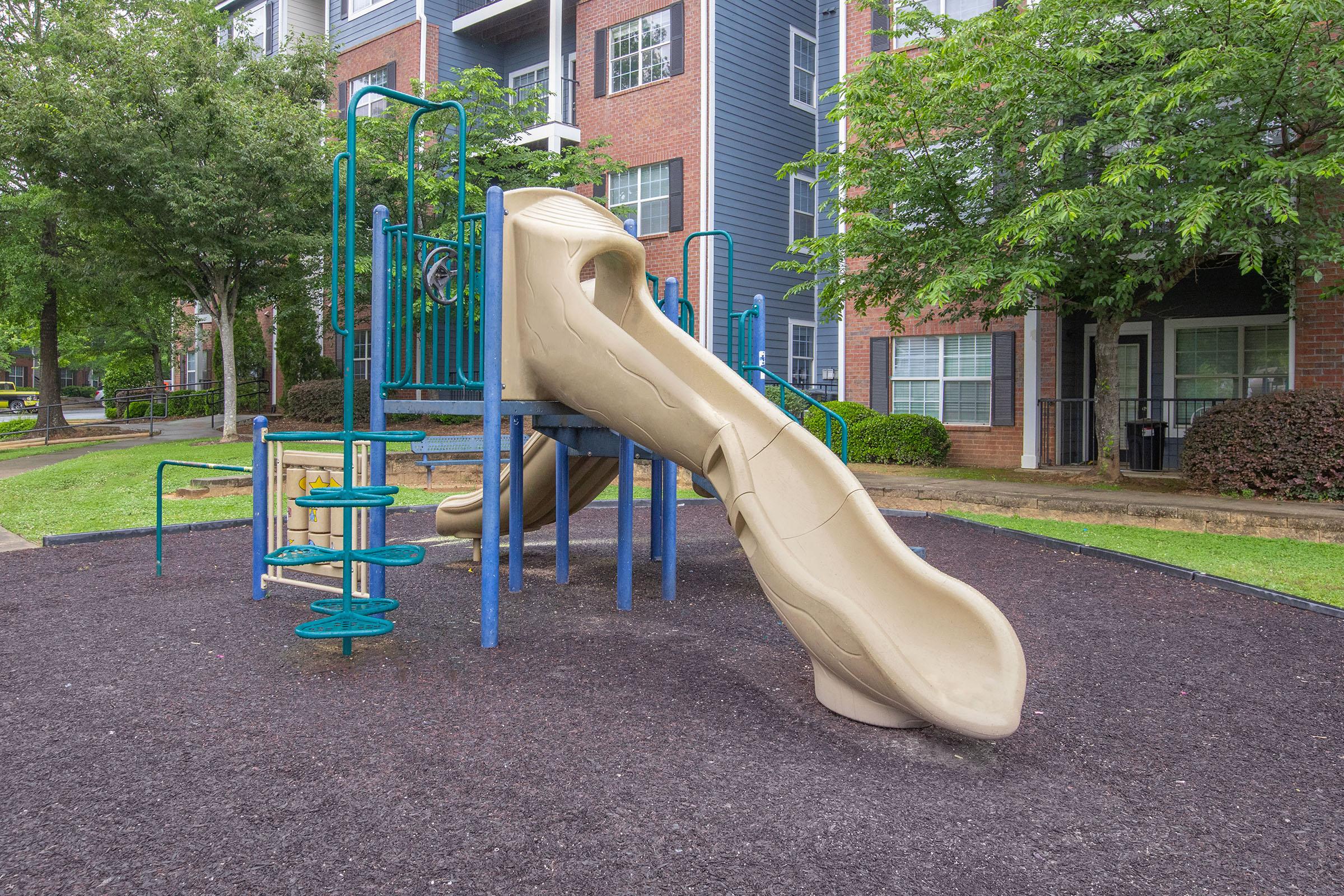 A playground structure featuring a large beige slide, climbing equipment, and surrounding grassy areas, set against a backdrop of residential buildings.