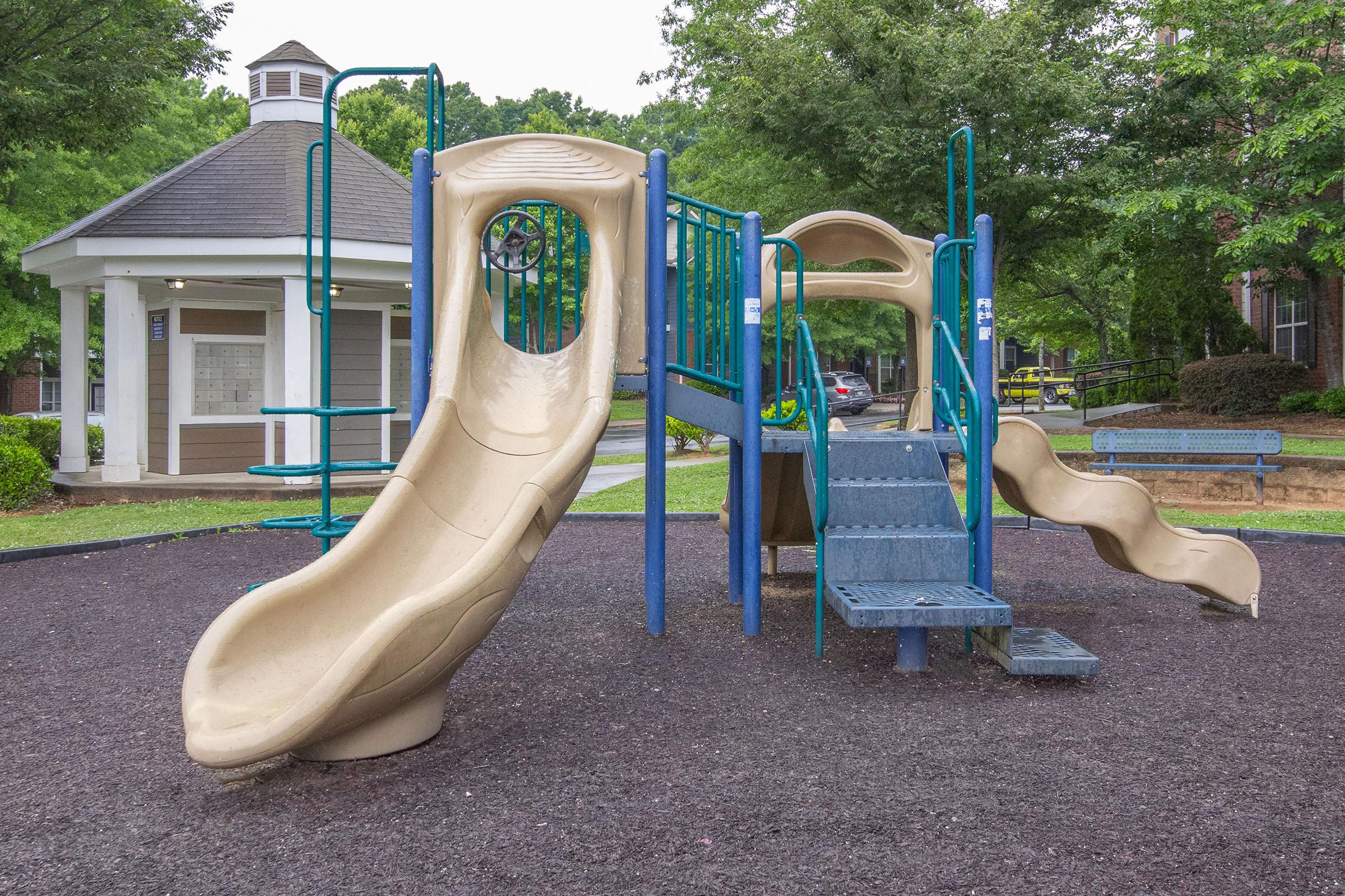 Playground structure featuring two slides, climbing steps, and a circular cutout, situated on a rubberized surface with surrounding greenery and a gazebo in the background.