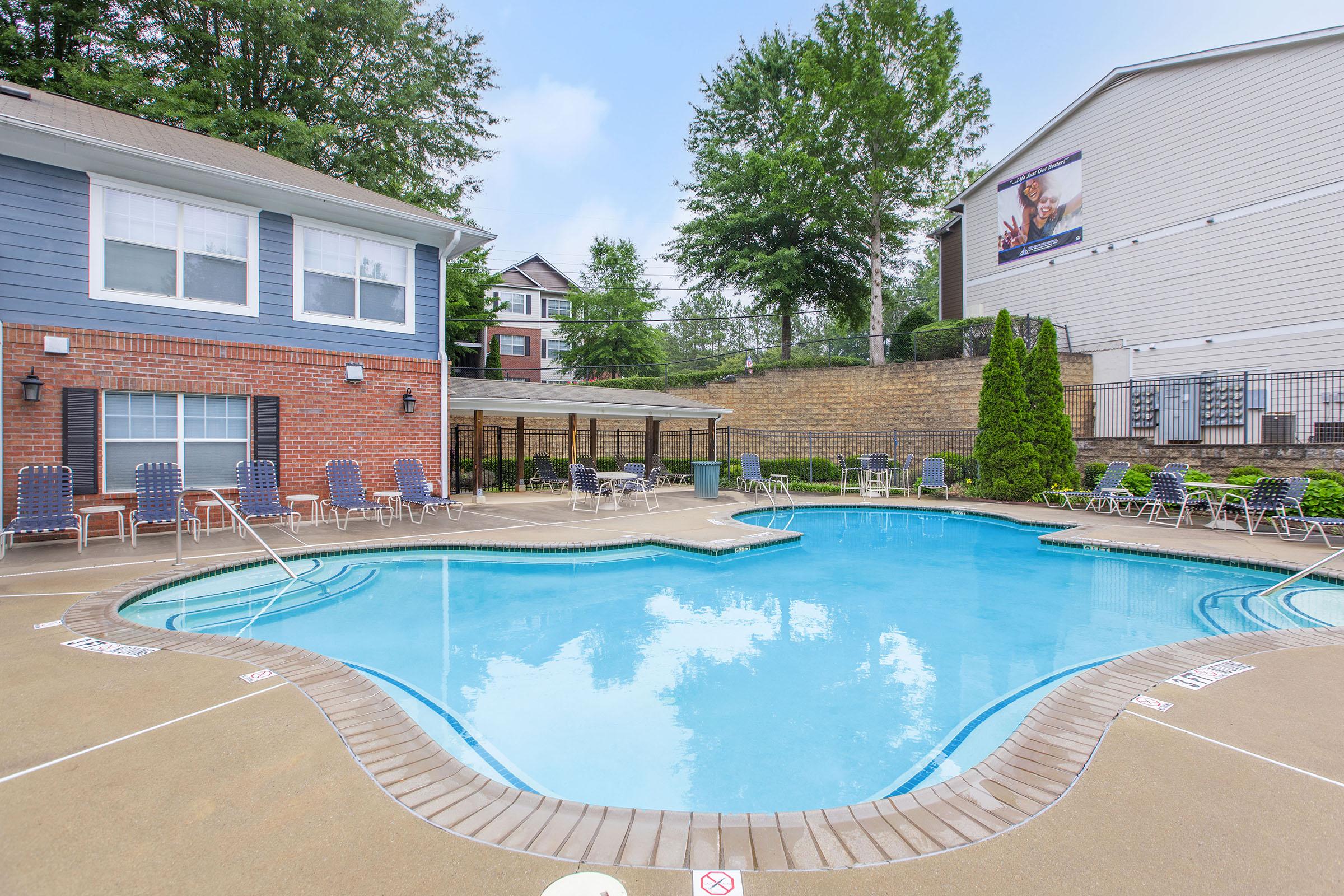 A swimming pool with a unique shape, surrounded by lounge chairs and shaded areas. The pool area is adjacent to a residential building with a blue and brick exterior and greenery in the background.