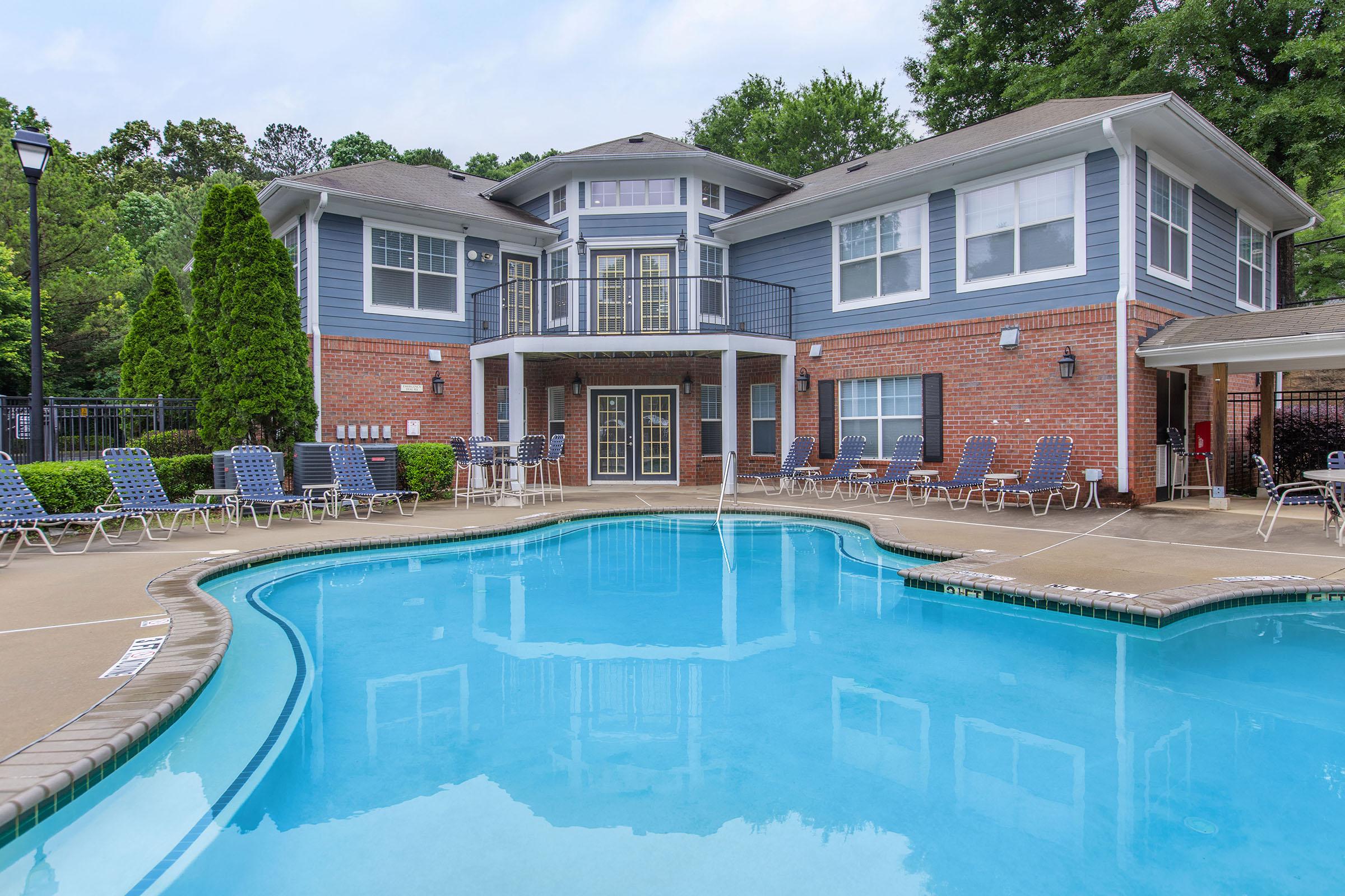 A residential building with blue and brick exterior surrounding a swimming pool. Lounge chairs are lined up beside the pool, and trees are visible in the background. The sky is partly cloudy.