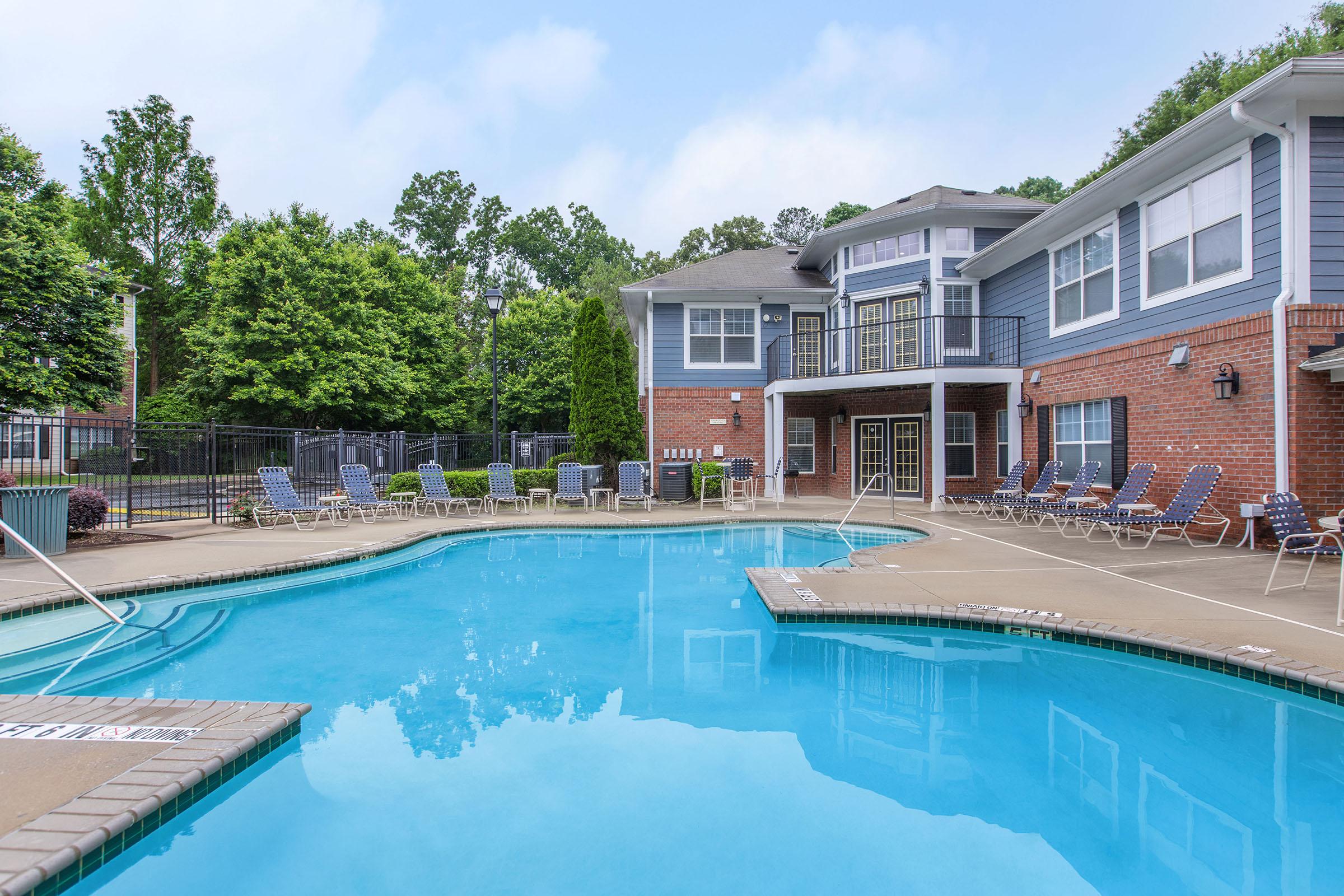 A clear blue swimming pool surrounded by a patio area with lounge chairs. The pool is framed by a brick building with balconies and large windows, set among green trees and landscaped areas.