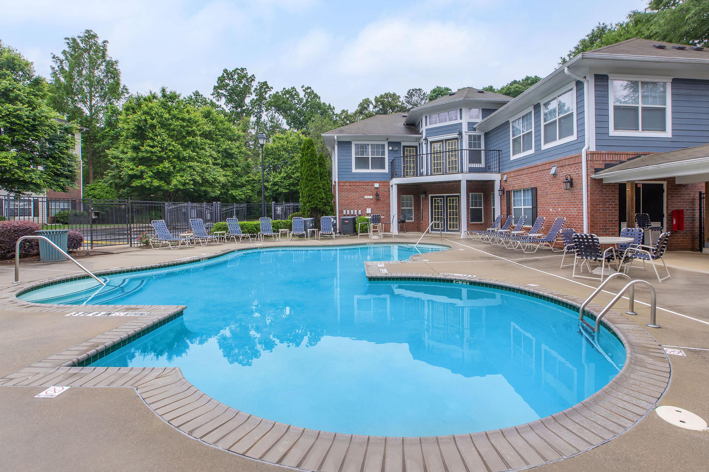 A clear blue swimming pool surrounded by lounge chairs, with a brick building and green trees in the background.