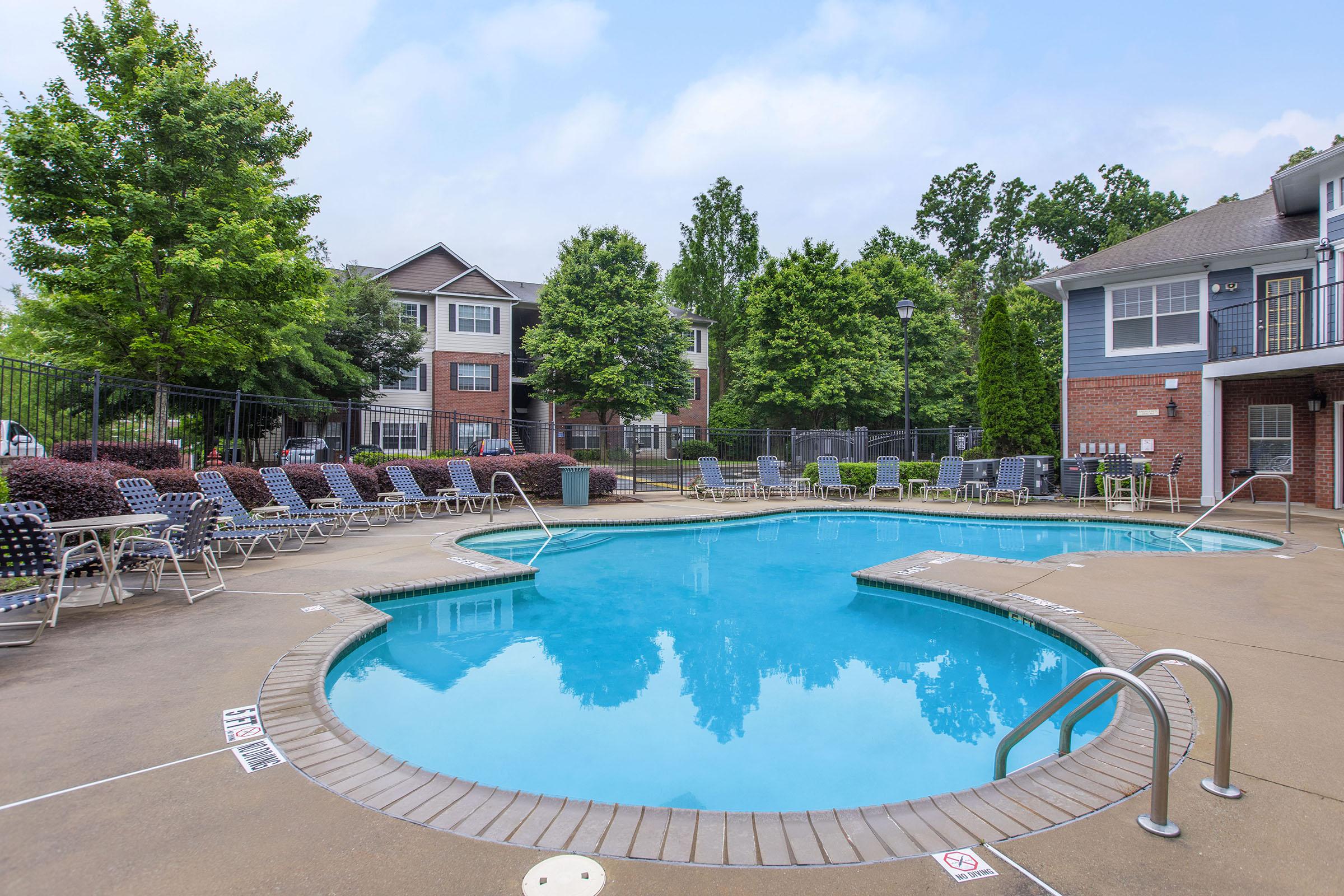 A sunny outdoor swimming pool area with clear blue water, surrounded by lounge chairs and neatly trimmed trees. Townhouse-style buildings are visible in the background.