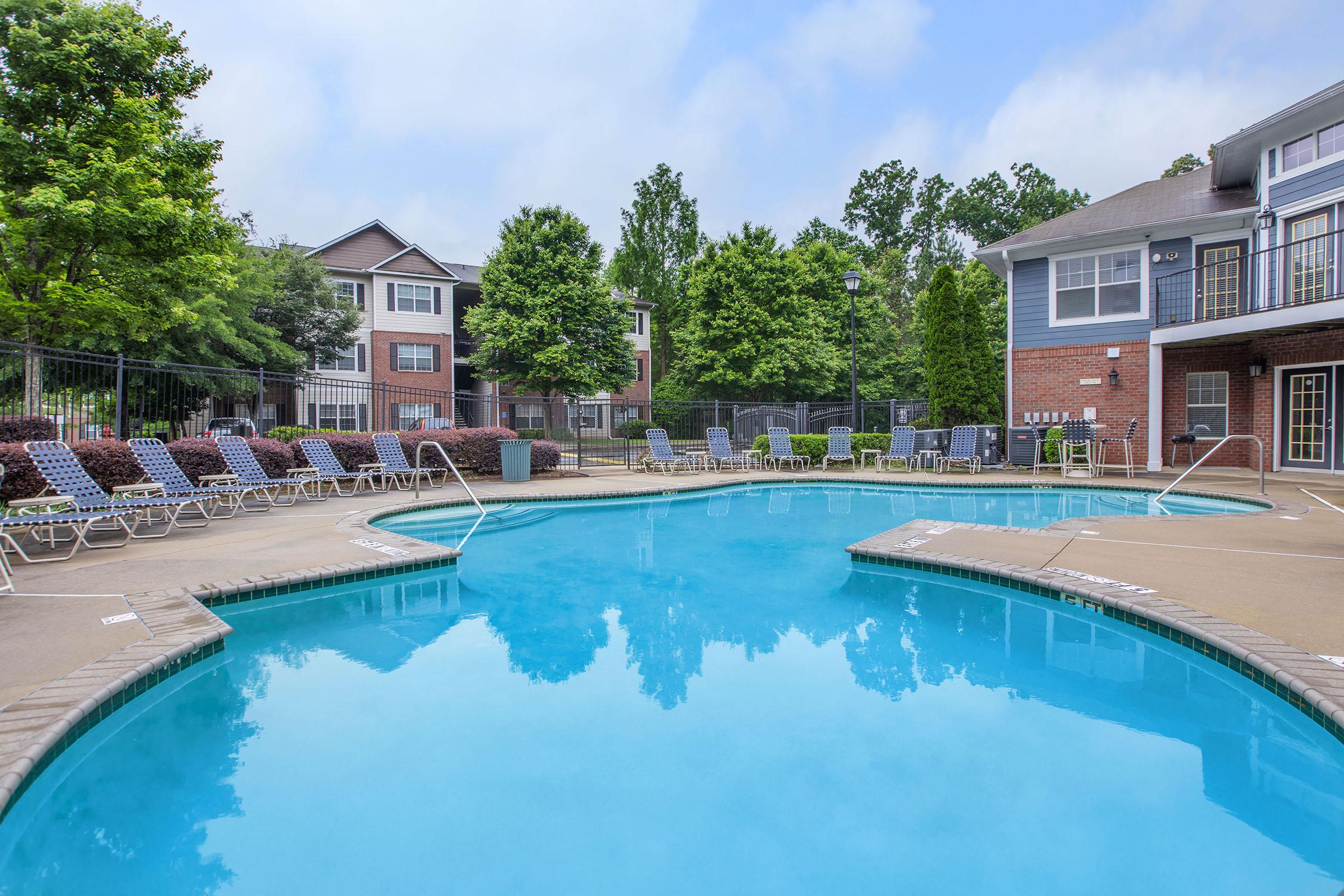 A clear blue swimming pool surrounded by lounge chairs, with trees and apartment buildings in the background. The scene is bright and inviting, perfect for relaxation.