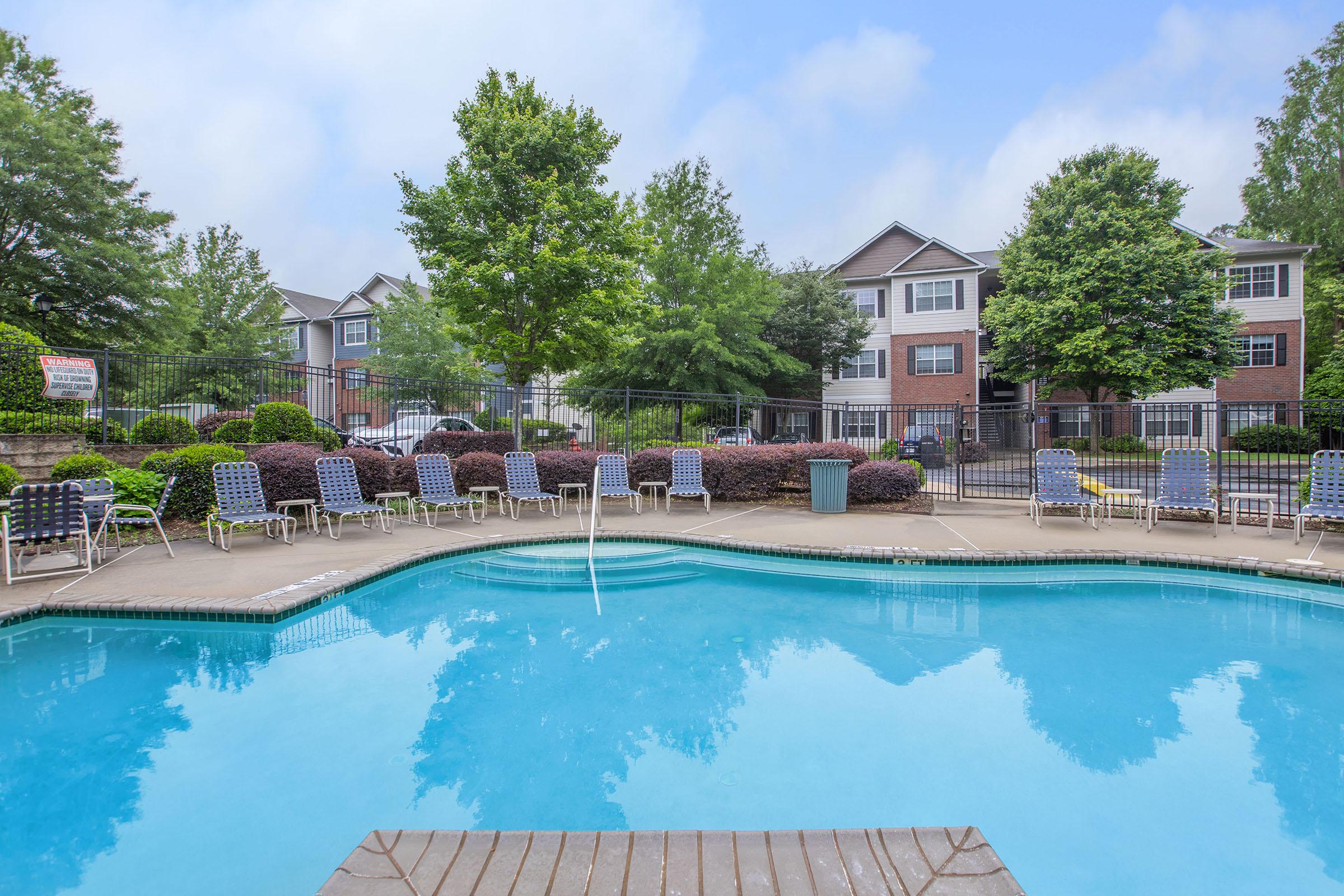 A view of a swimming pool surrounded by lounge chairs, with a green landscape of trees and apartment buildings in the background. The water is clear and inviting, and there are no people present.