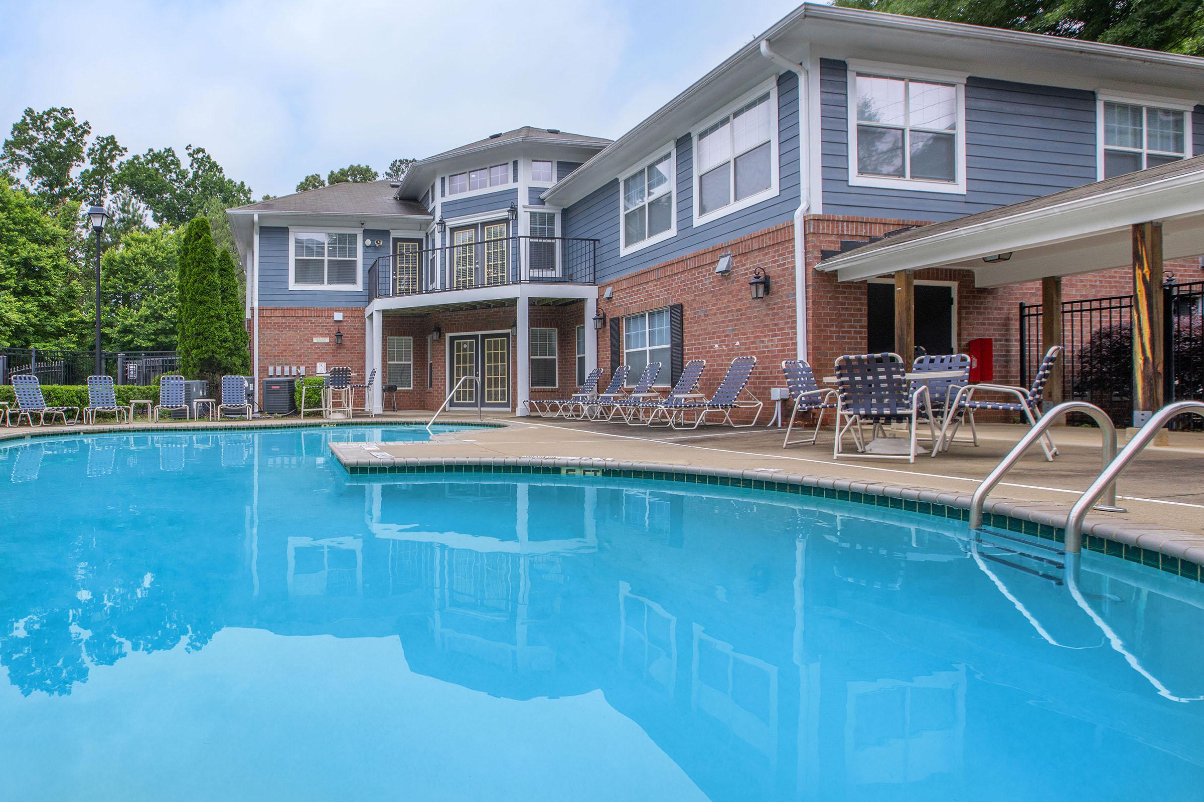 Swimming pool area featuring a clear blue pool with a surrounded patio, lounge chairs, and a residential building in the background.