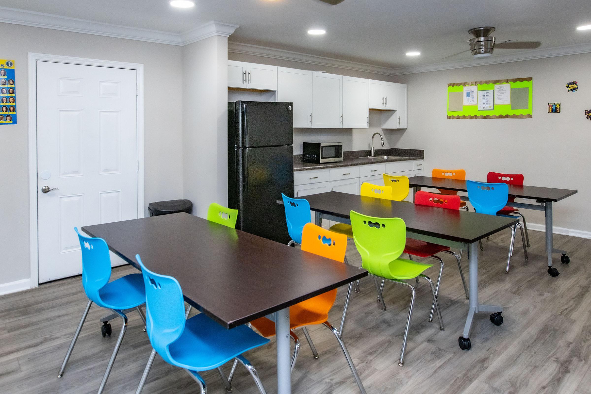 A bright and modern classroom or dining area featuring multiple tables with colorful chairs in blue, red, orange, and green. The walls are light-colored, and there are white cabinets, a black refrigerator, and a microwave visible. A bulletin board with green trim is mounted on one wall, and there is a door leading to another space. The flooring is a light wood laminate.