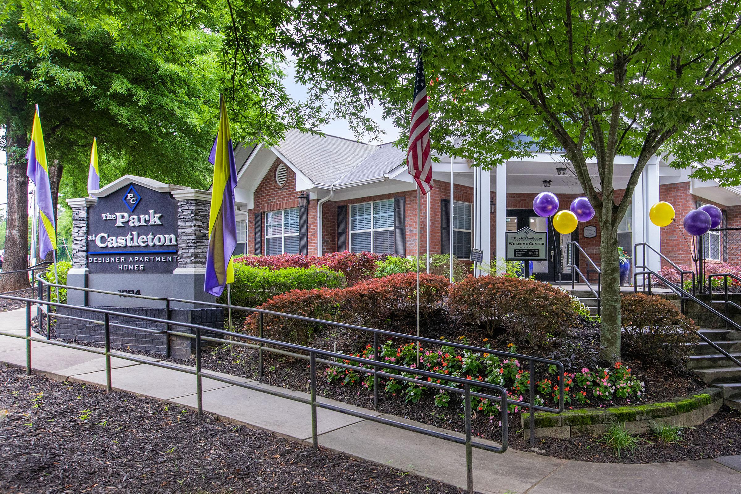 A landscaped entrance to an apartment community named "The Park Castleton," featuring a stone sign, decorative flags, colorful balloons, and well-maintained shrubs and flowers. The building has a welcoming facade with a canopy and steps leading to the entrance.