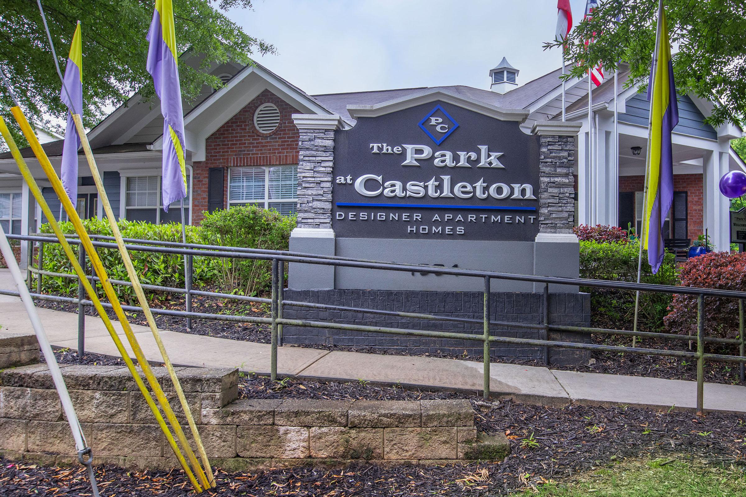 Sign for "The Park at Castleton" featuring the words "Designer Apartment Homes," with a landscaped entrance and colorful flags visible.