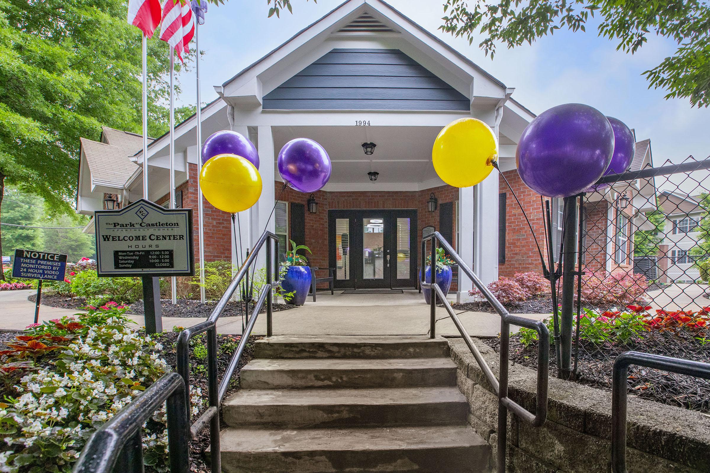 A welcoming entrance to a building with stairs leading up to the door. The steps are flanked by potted plants and colorful balloons in purple and yellow, creating a festive atmosphere. Flags are visible in the background, and a sign indicates the presence of a welcome center.