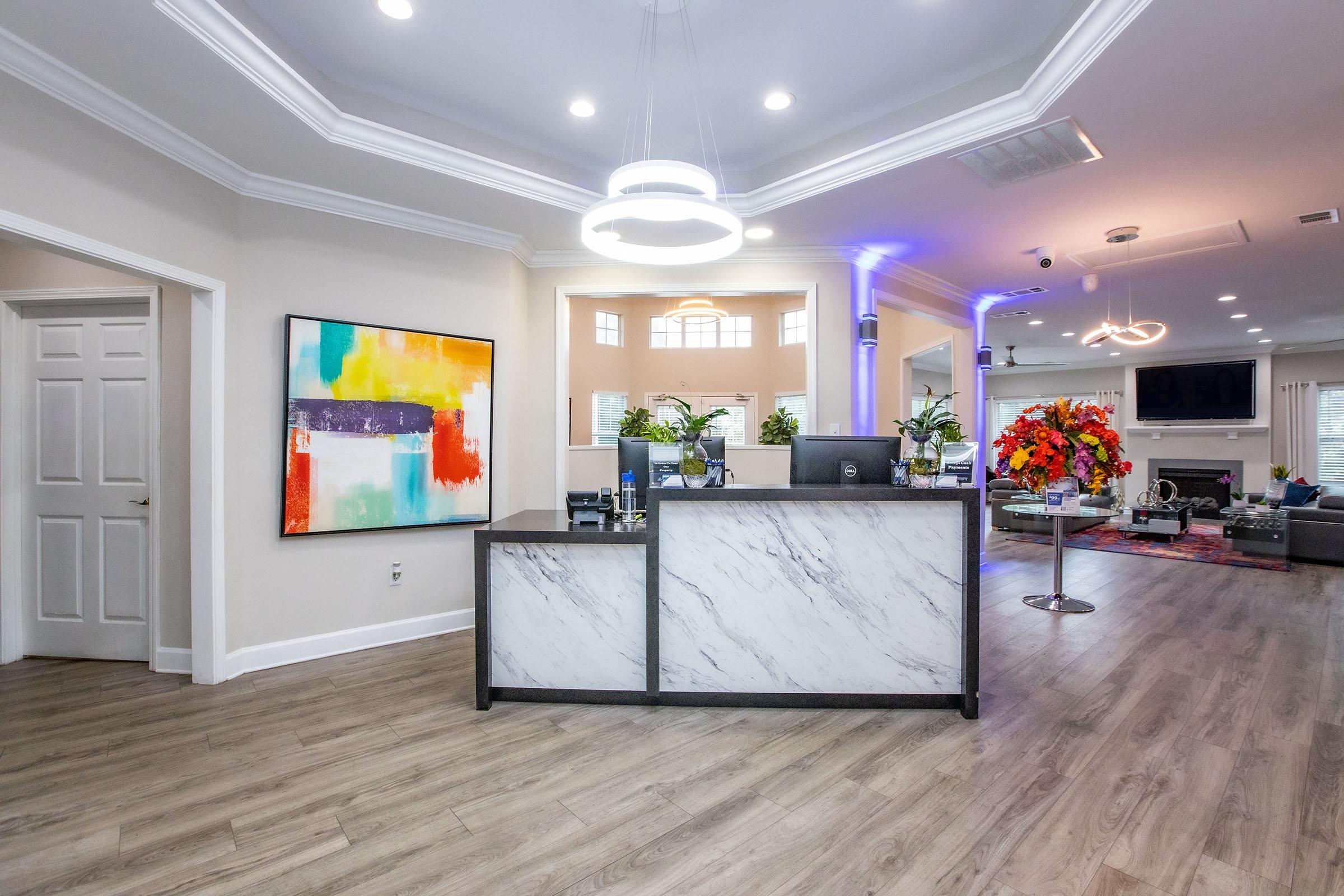 A modern reception area featuring a marble-front desk, colorful abstract artwork on the wall, and vibrant floral arrangements. The space is well-lit with a contemporary light fixture and has a neutral color palette with wooden flooring. A doorway is visible on the left, and a spacious lounge area in the background is partially visible.