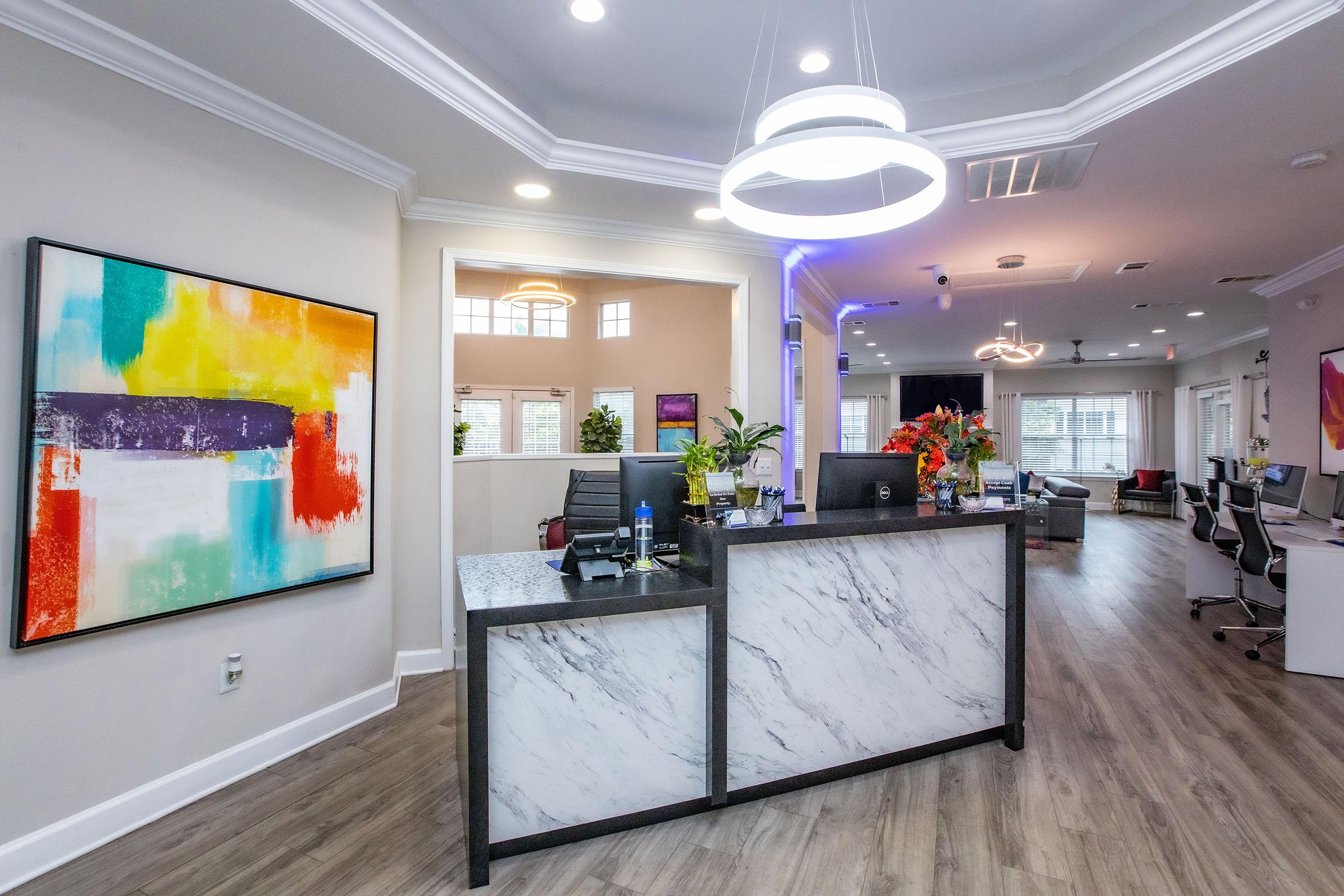 Modern reception area with a stylish marble front desk, vibrant artwork on the wall, and well-lit space featuring potted plants. The background includes a seating area and workstations, creating a welcoming atmosphere.