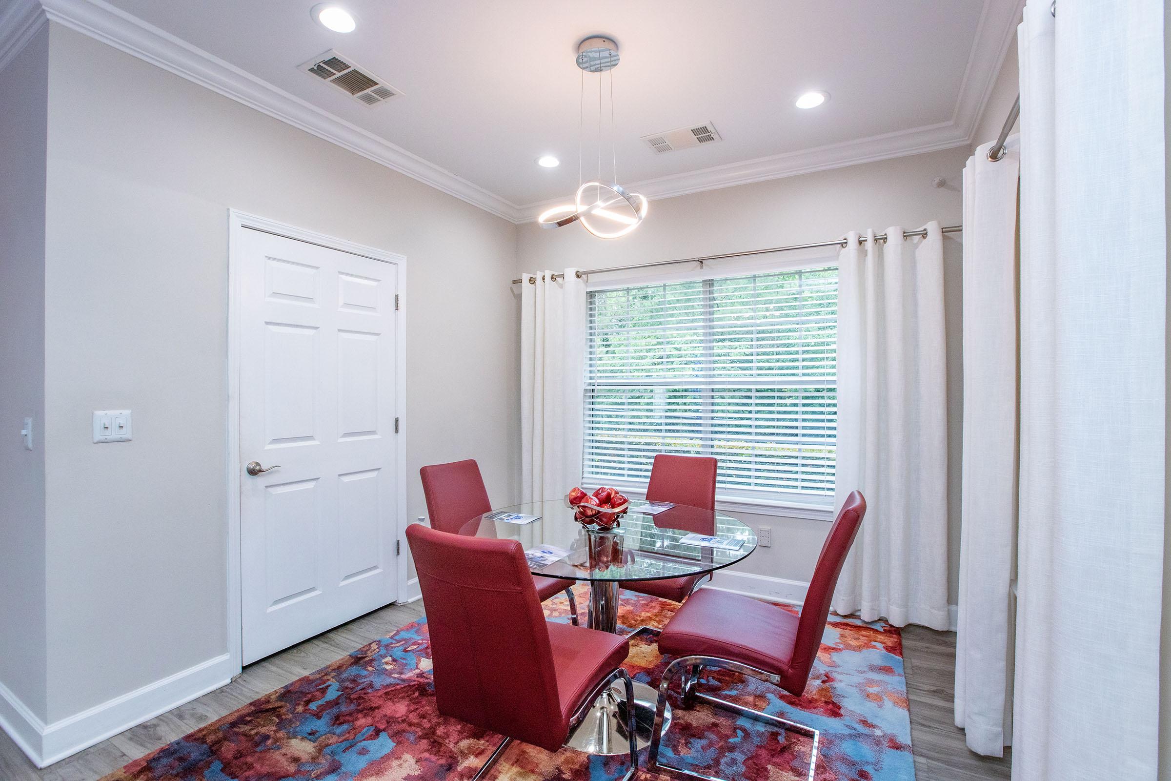 A modern dining area featuring a round glass table with four red chairs. The space has a colorful rug beneath the table, a window with white curtains, and soft overhead lighting. A door is visible on one side of the room.