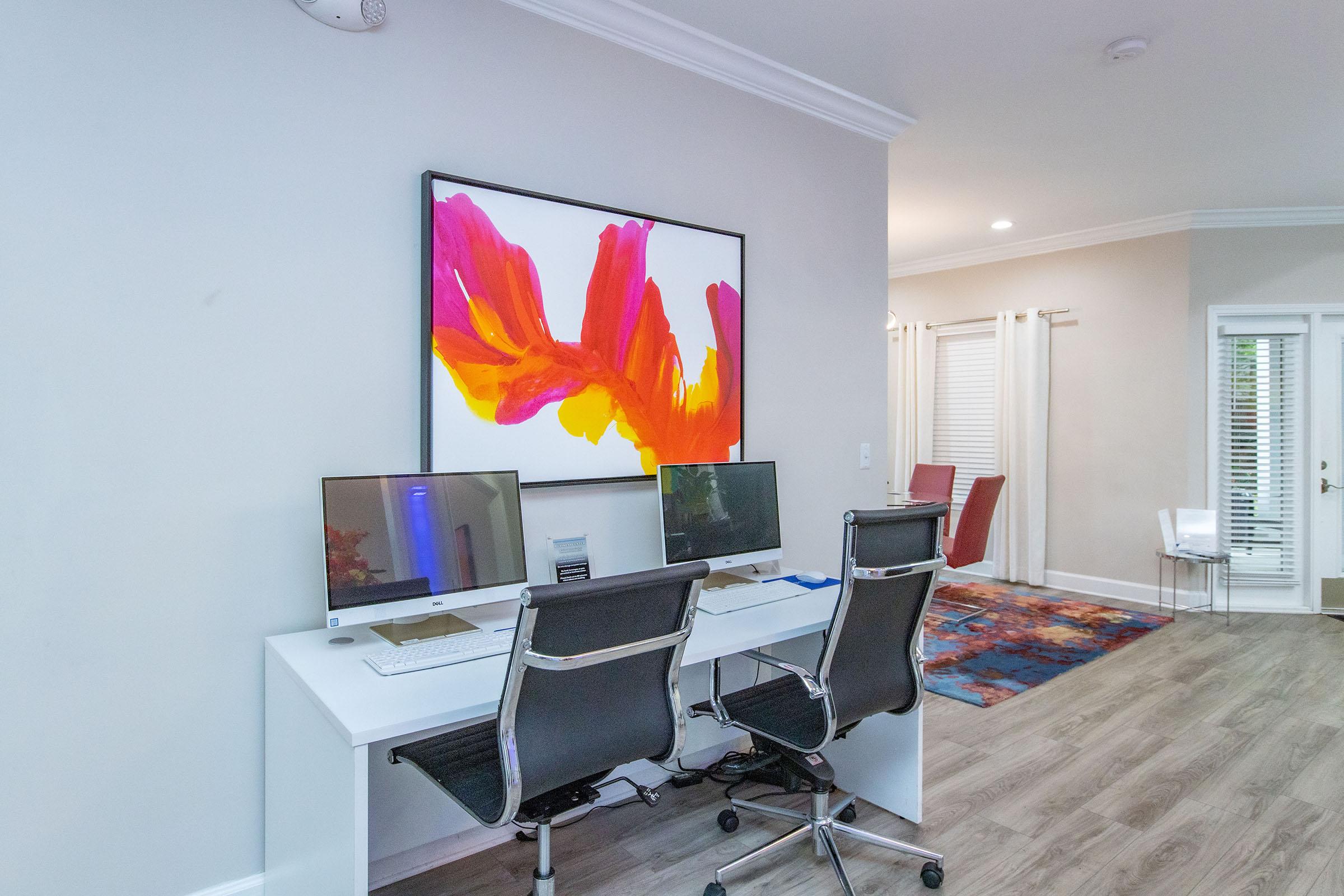 A modern home office featuring two black ergonomic chairs at a white desk with two computer monitors. A colorful abstract painting in shades of pink, orange, and yellow is displayed on the wall above the desk. In the background, there is a small dining area with a red chair and large windows with sheer curtains. The flooring is light wood.