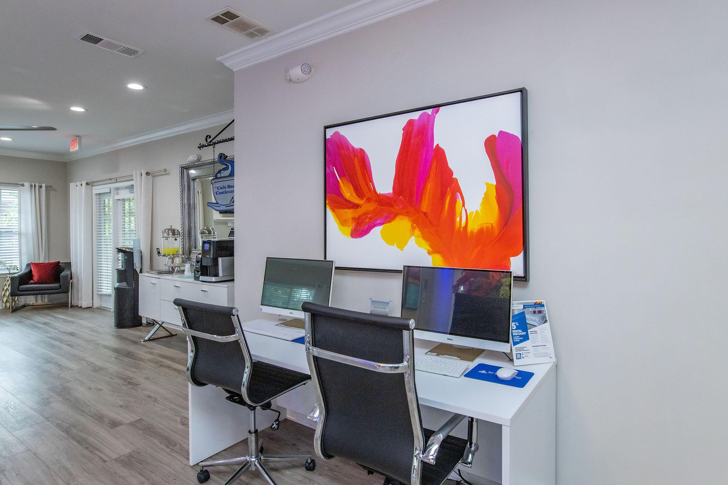 A modern workspace featuring two computer desks with monitors, a colorful abstract painting hanging on the wall, and a cozy seating area in the background. The room has light-colored walls and wooden floors, creating a bright and inviting atmosphere.