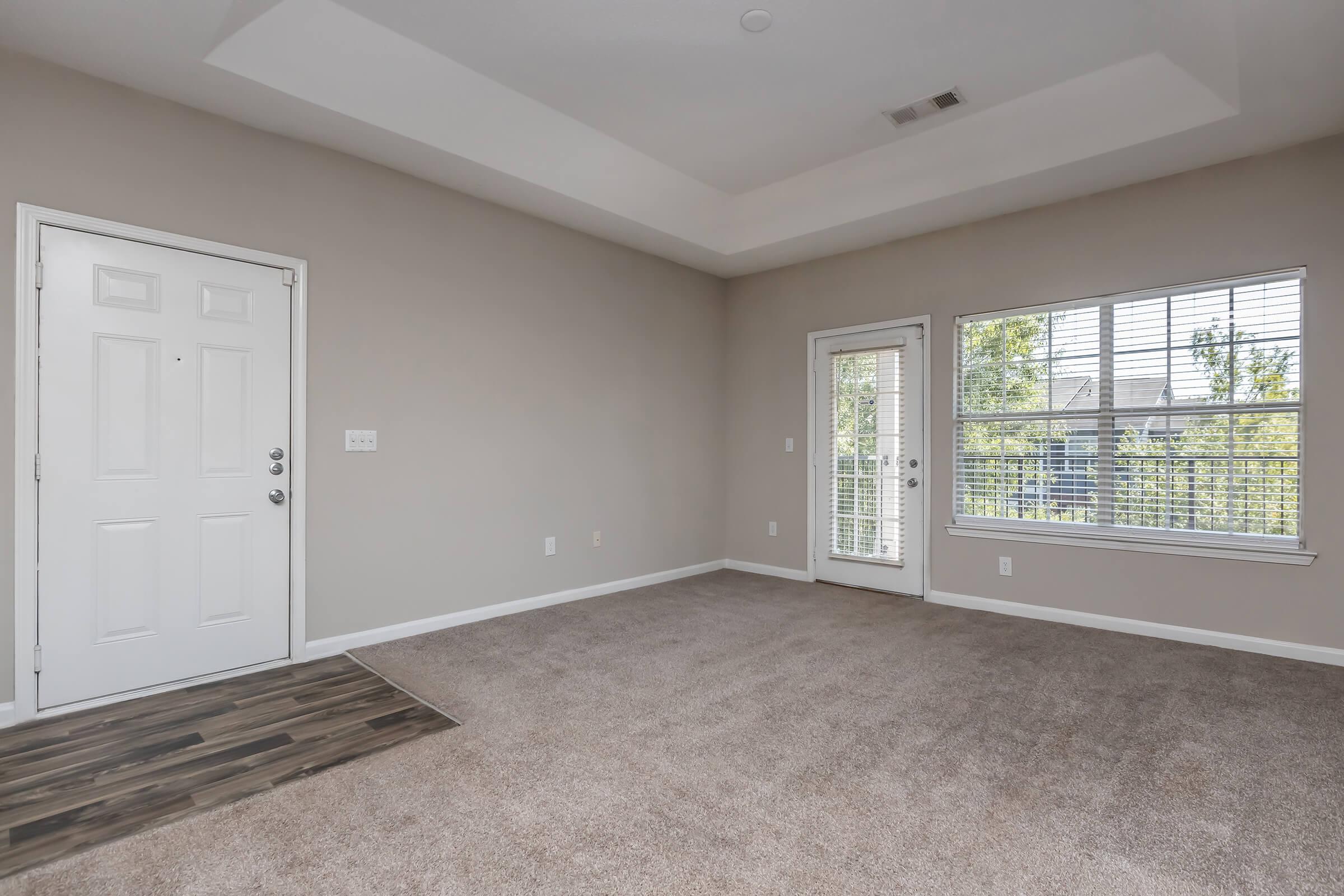 A spacious, empty living room featuring light gray walls and a carpeted floor. There is a front door on the left and a set of double doors leading to a balcony on the right, with large windows allowing natural light to enter. The room has a ceiling with a subtle architectural detail.