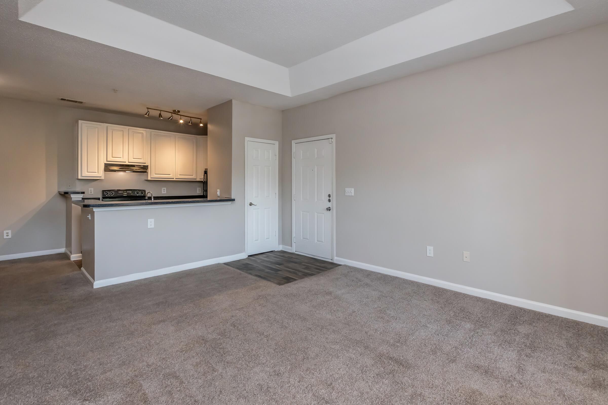 A spacious, empty living area with light beige walls and grey carpet. The room features a small entryway with a door leading outside. A kitchen area with white cabinets and modern appliances is visible in the background, partially separated by a breakfast bar. Natural light comes from an overhead fixture.