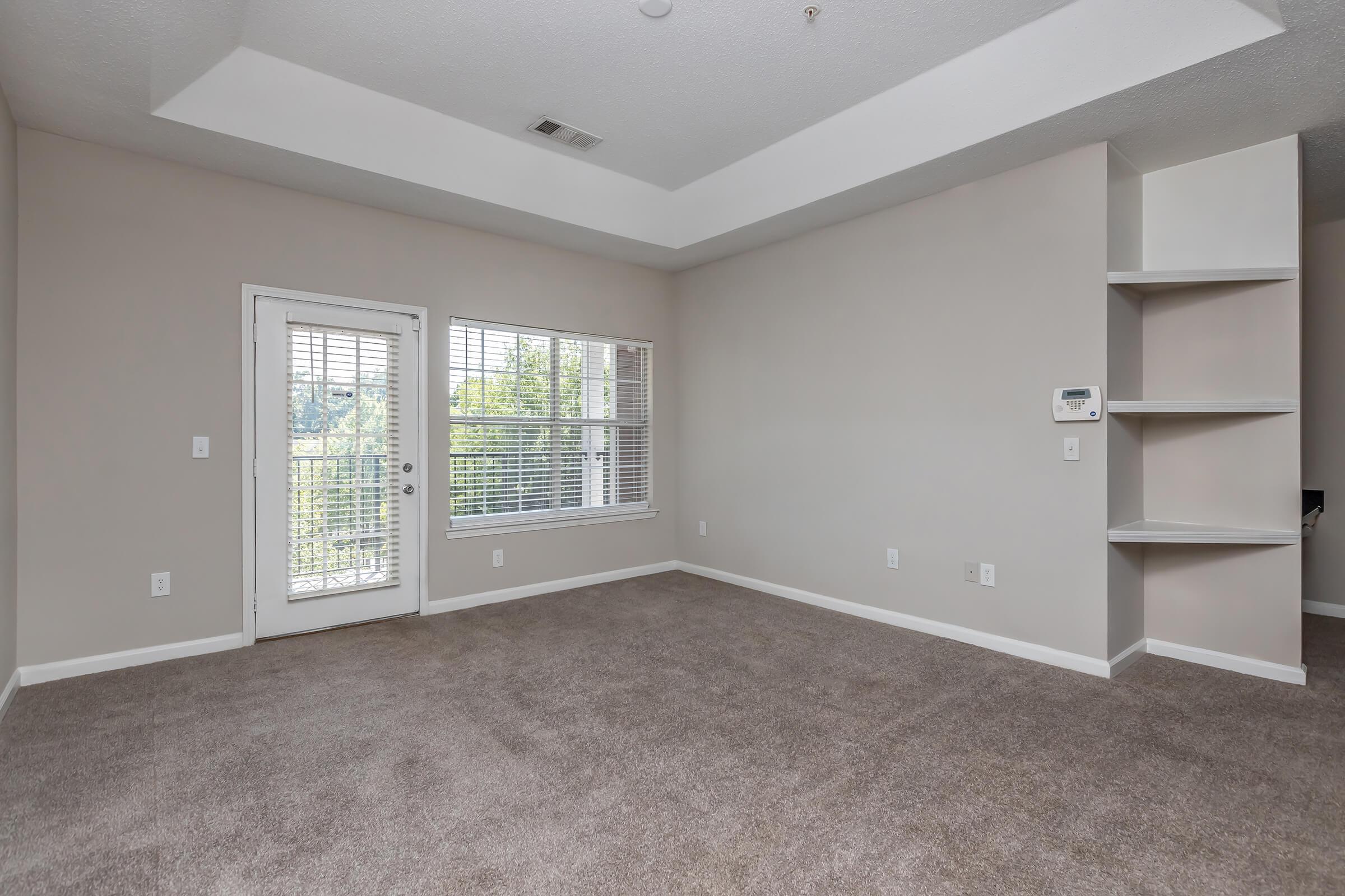 Spacious, empty room with light beige walls and carpeted flooring. A sliding glass door leads to a balcony, and a large window offers natural light. Built-in shelves are present on one wall, and there is a small thermostat on another. The ceiling features a slight indentation for added architectural detail.