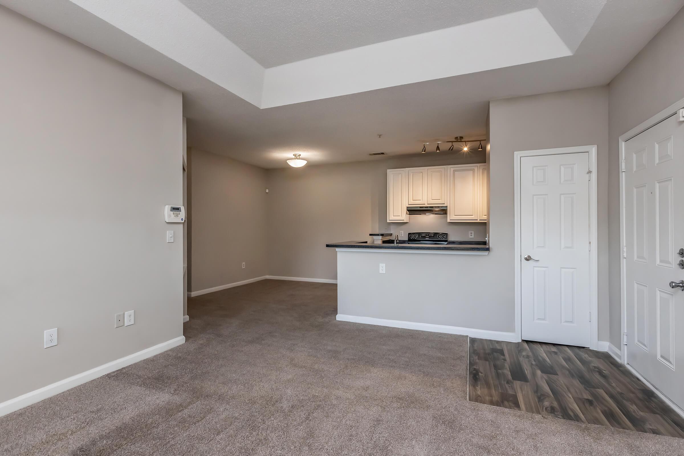 A spacious, unfurnished living area featuring beige carpet and light gray walls. The kitchen is partially visible, equipped with white cabinets and a countertop. A door leads to the outside, illuminating the room with natural light. The layout is open and inviting, suitable for various interior designs.