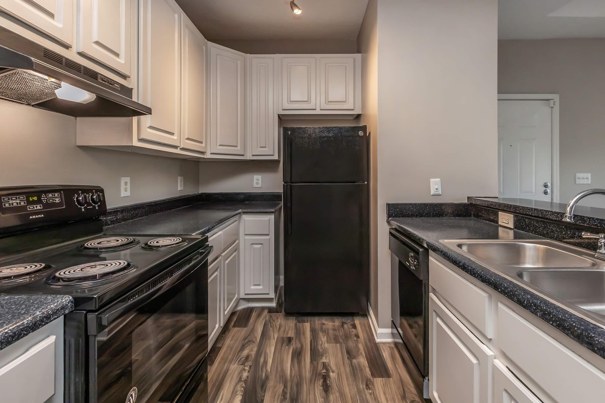 A modern kitchen featuring dark countertops, white cabinetry, and stainless steel appliances. The layout includes a black refrigerator, an oven, and a dual-basin sink, with wood-look flooring and neutral-colored walls creating a bright and inviting atmosphere.