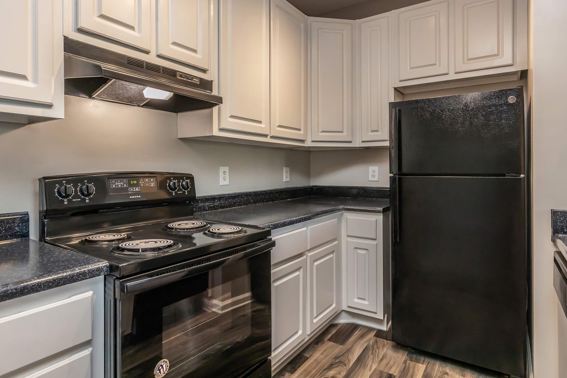 A modern kitchen featuring dark countertops, white cabinets, and a black refrigerator. The kitchen includes a black electric stovetop with an oven below, a range hood above, and ample cabinet space for storage. The flooring is dark, adding contrast to the light-colored cabinetry.