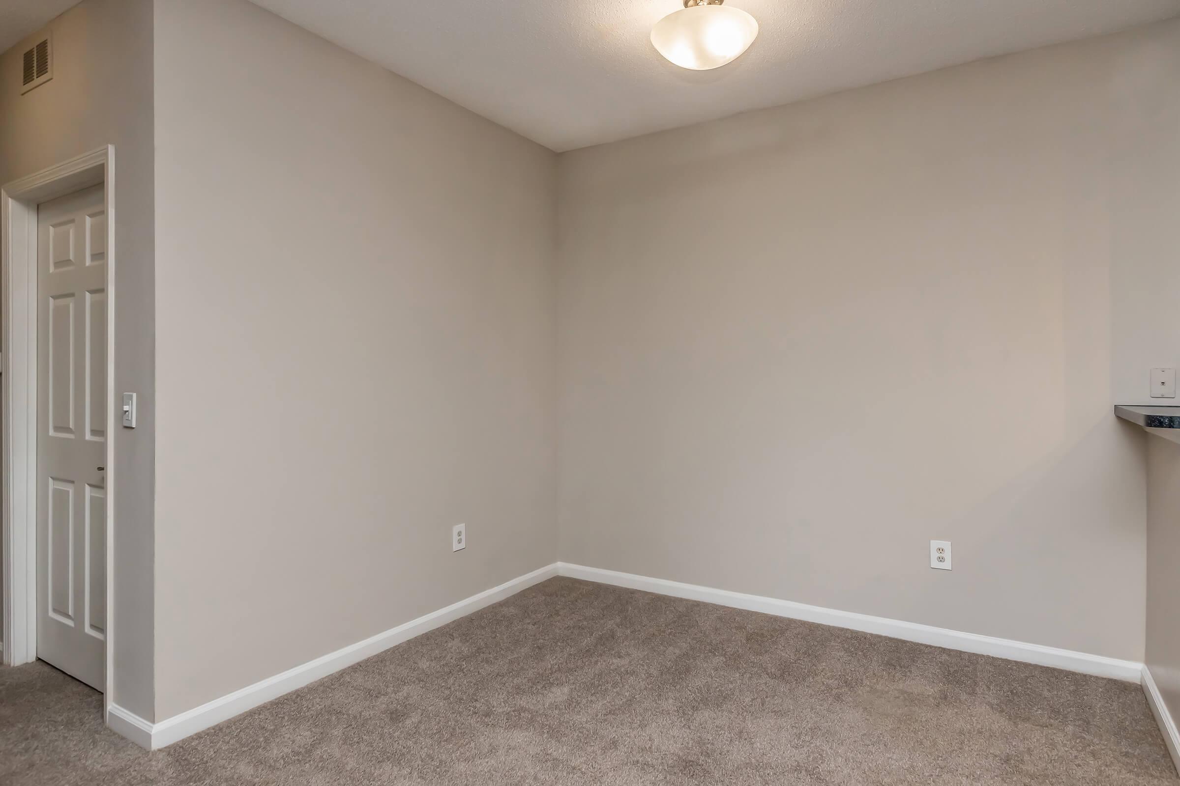 A vacant room with light gray walls and plush beige carpet. The space features a ceiling light fixture and a white door on the left. A wall on the right displays an area where a shelf or counter may be mounted. The room is bright and unoccupied, showcasing a minimalist and neutral design.