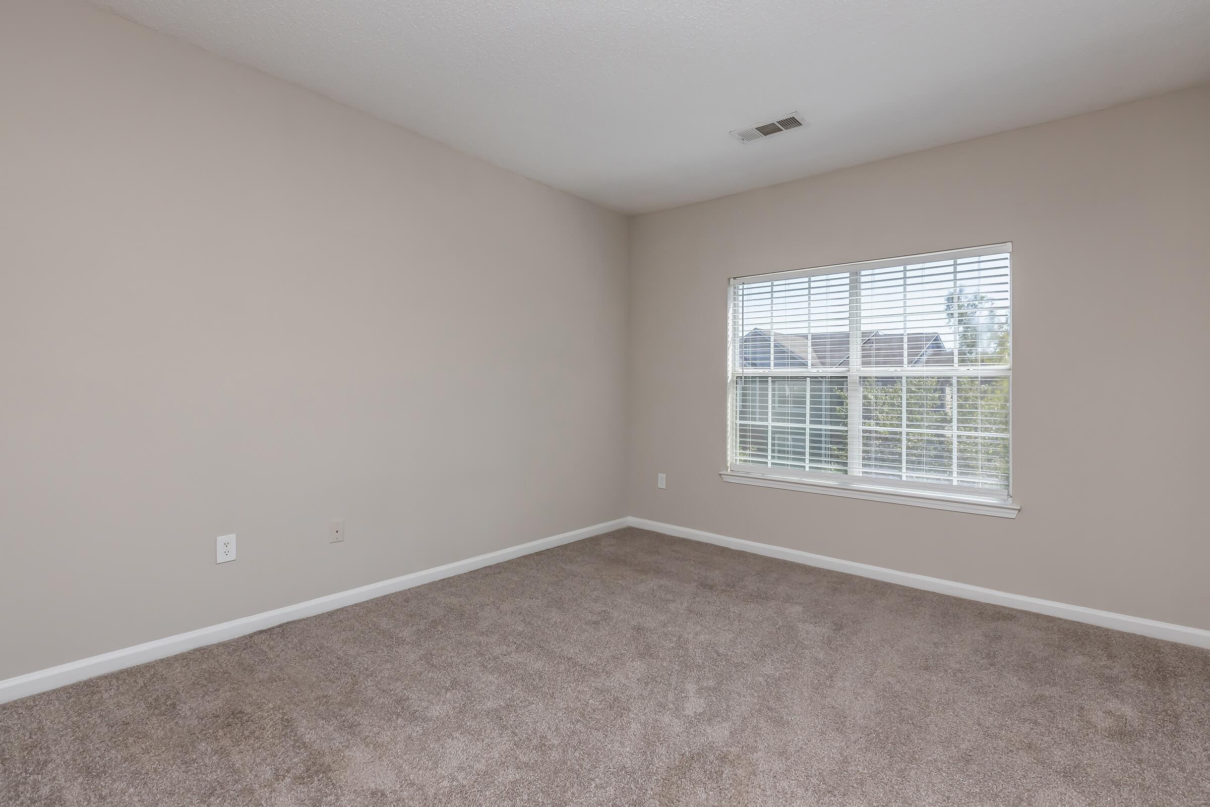 A spacious, empty room with light beige walls and soft, tan carpet. A single window with white blinds allows natural light to enter, illuminating the space. The room features no furniture, suggesting potential for customization.